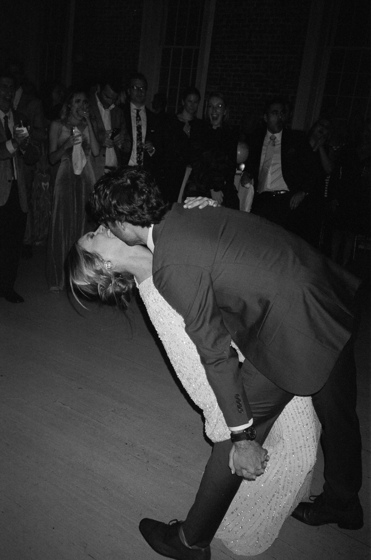Groom dipping bride on dance floor surrounded by wedding guests in dramatic black and white photo.