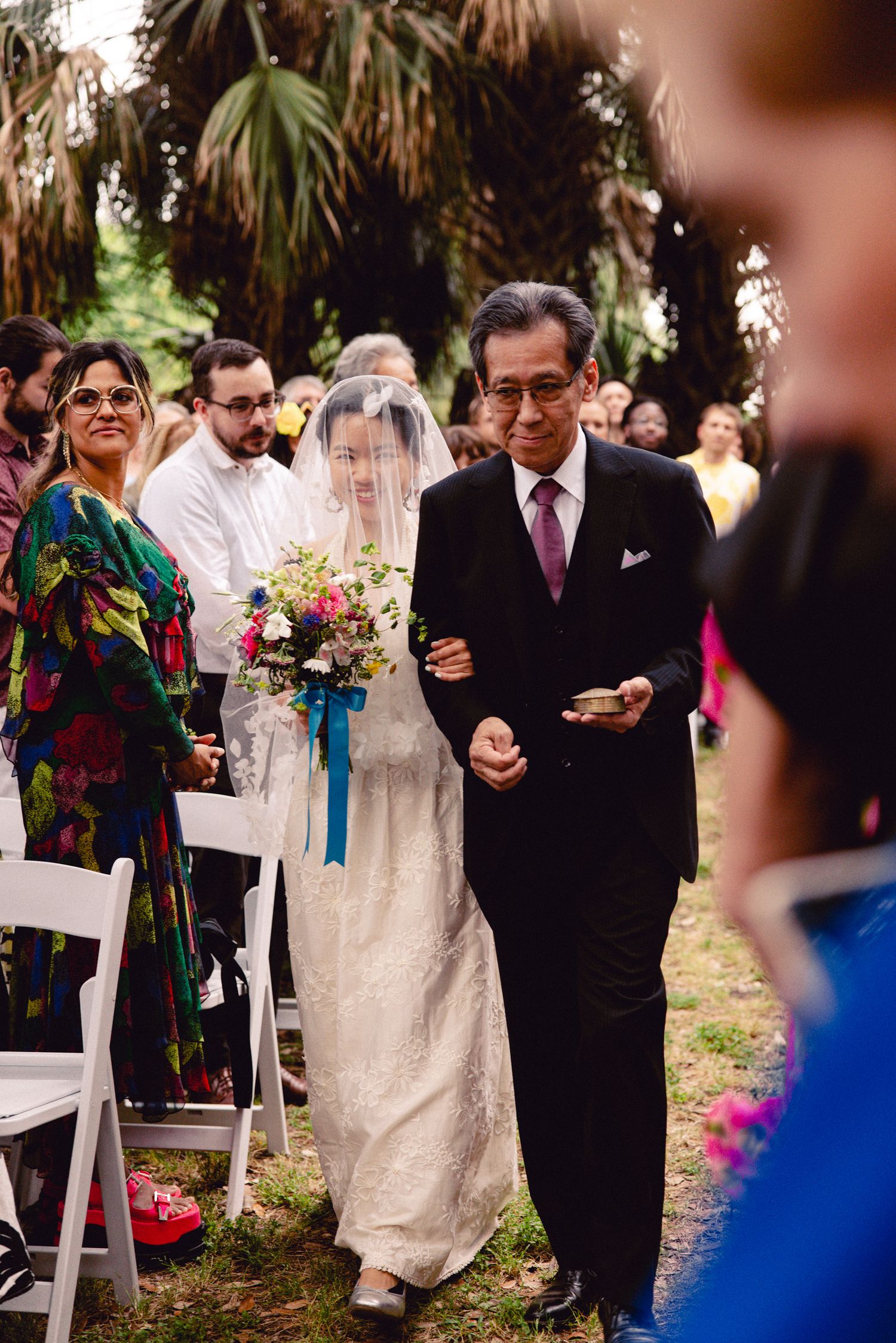 Father walking bride down the outdoor wedding aisle as guests watch from white chairs.