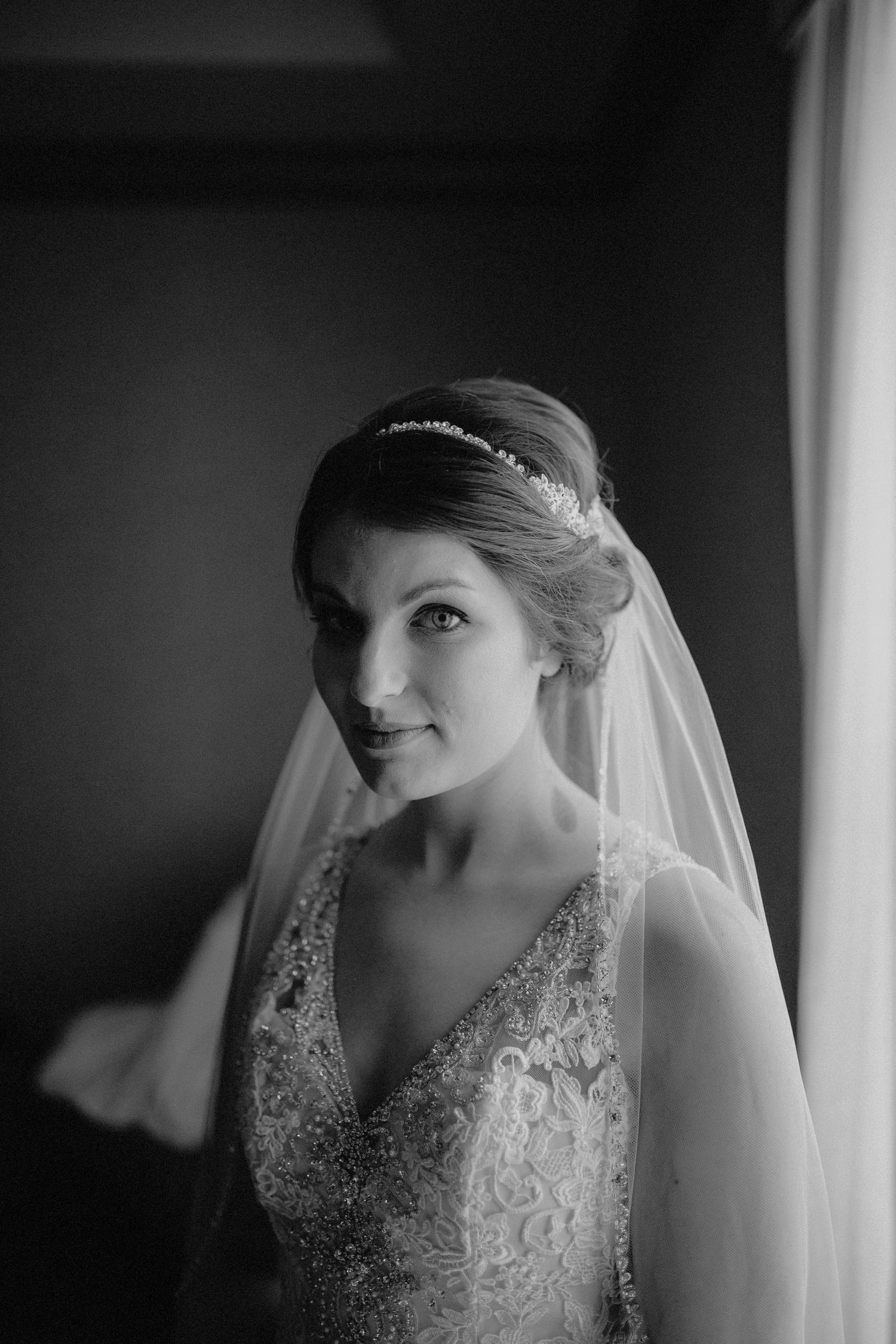 Elegant bride in lace gown and crystal tiara poses with veil in soft black and white portrait.
