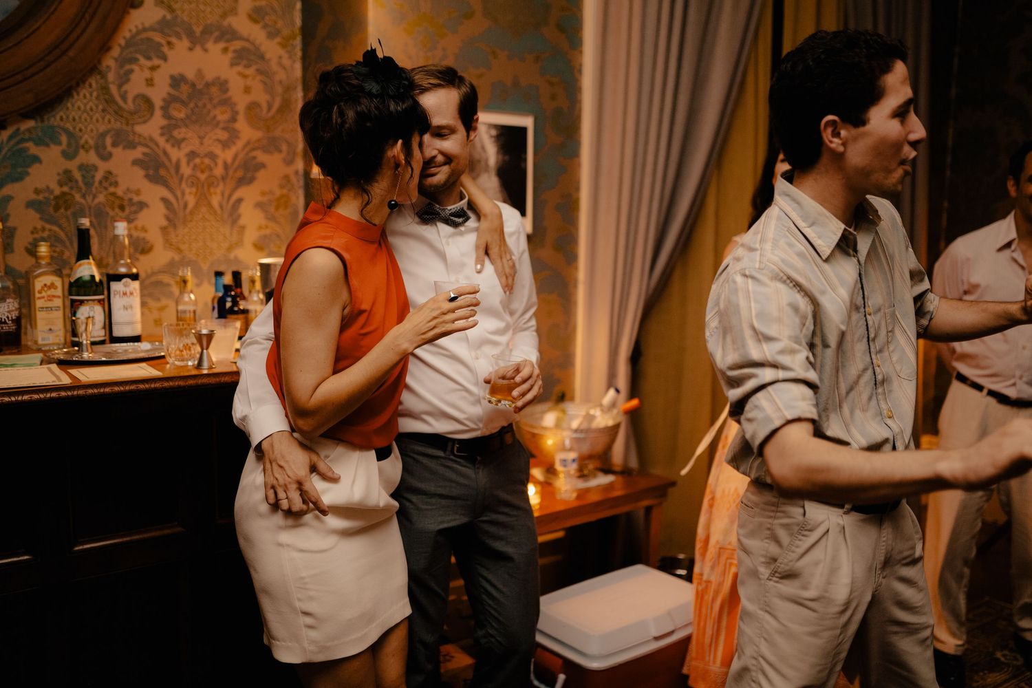 Guests laughing and socializing at a wedding reception near a bar with bottles displayed.