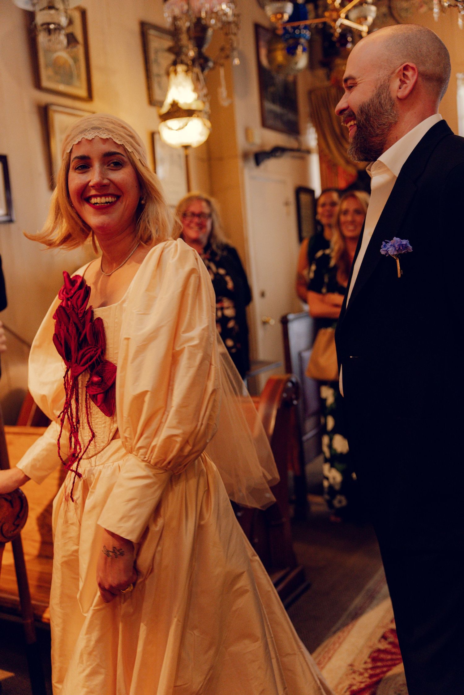 Bride in cream dress with red floral detail laughs with bald groom in dark suit at wedding ceremony.