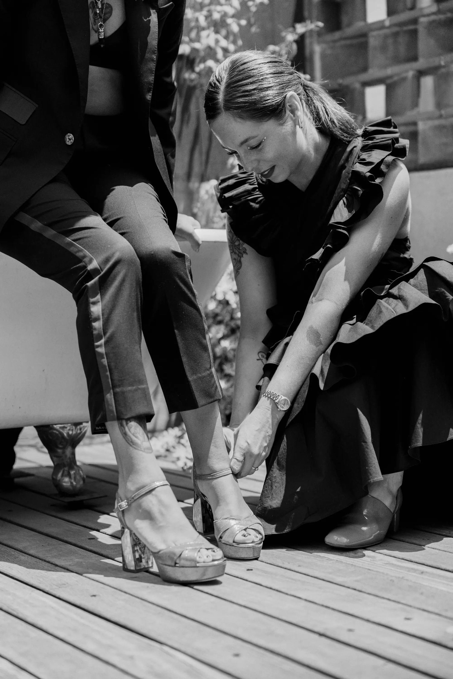 Bridesmaid in black dress helping bride with strappy heeled sandals on wooden deck.