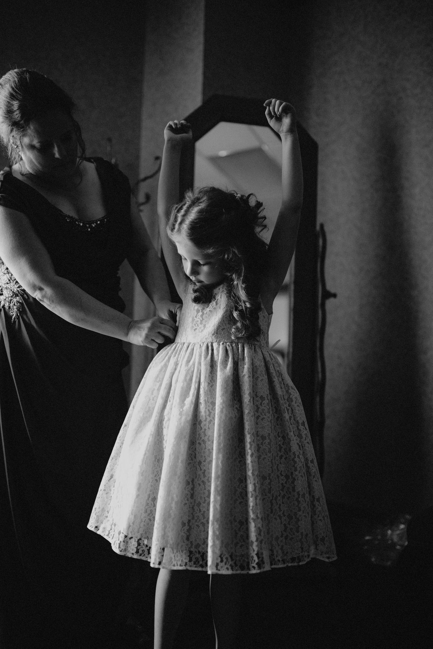 Woman helps young flower girl in white lace dress prepare in front of vintage mirror.