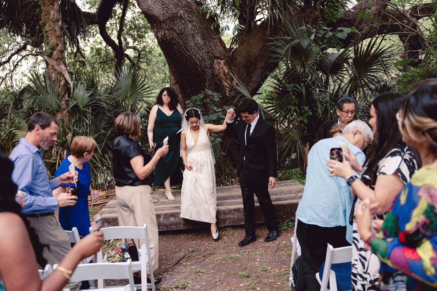 Bride and groom walk down the aisle as guests cheer and photograph at an outdoor wedding ceremony.