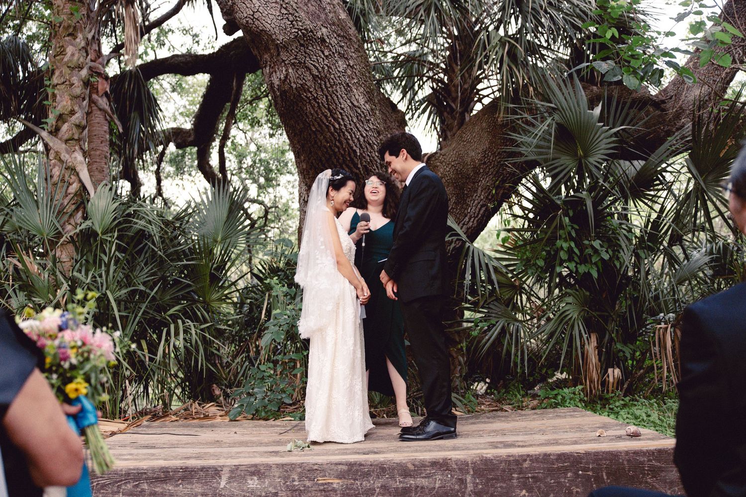 Bride and groom exchange vows under a large oak tree in a lush tropical garden ceremony.