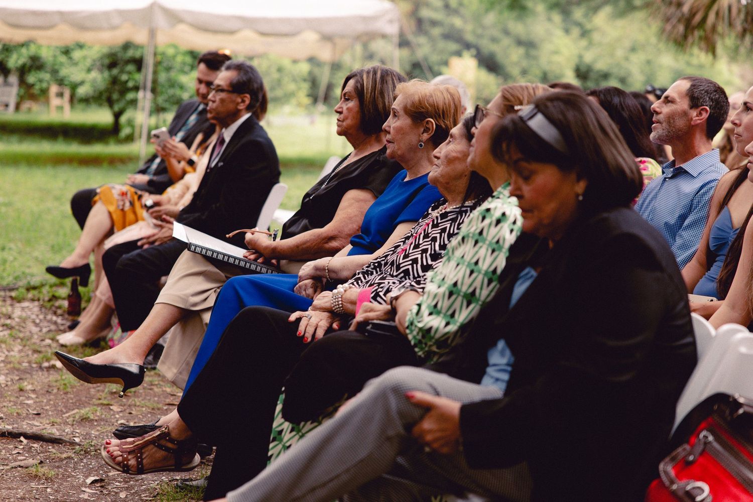 Diverse group of wedding guests seated outdoors on chairs, watching an outdoor garden ceremony.