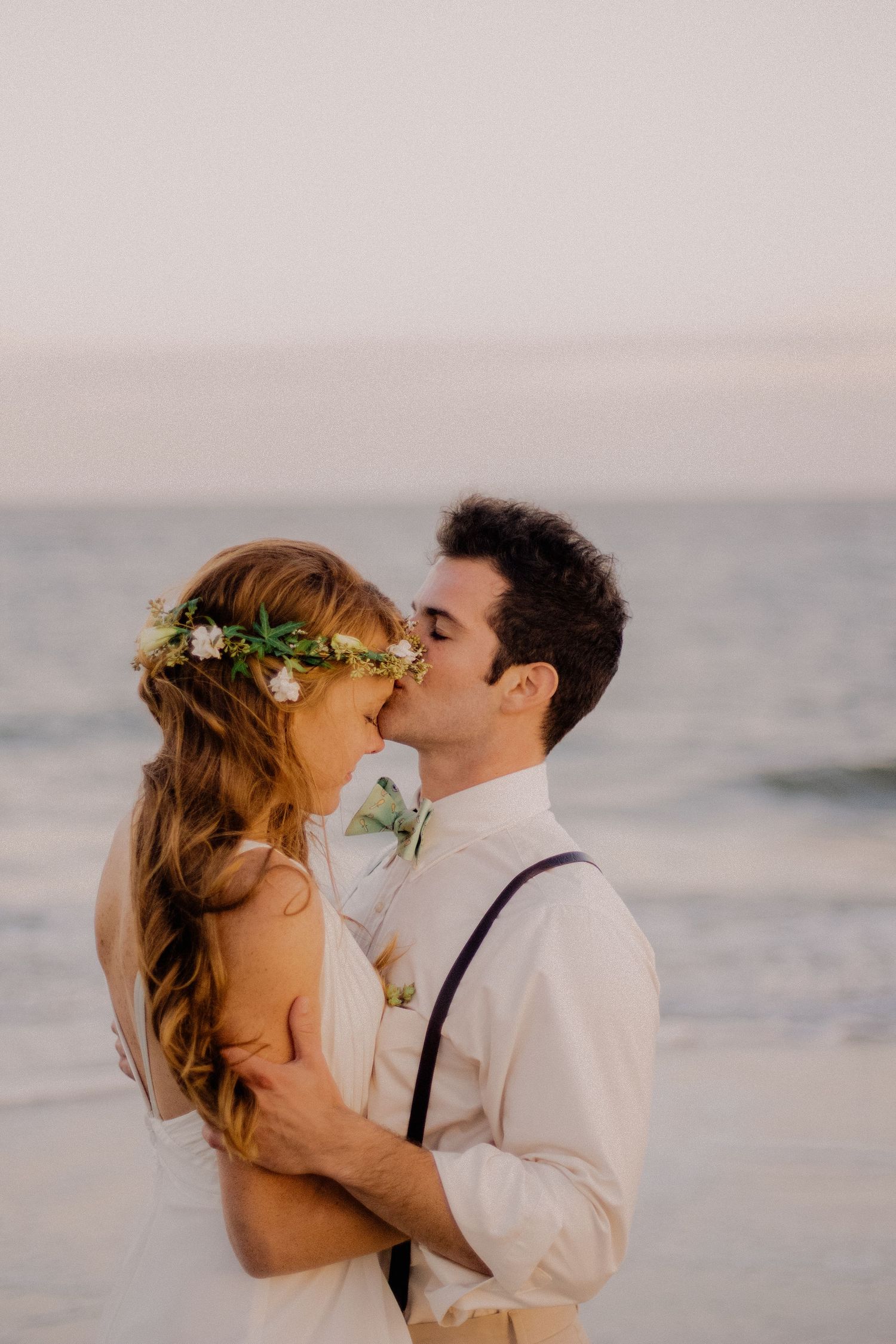 Groom kissing bride wearing floral crown on beach at sunset wedding.