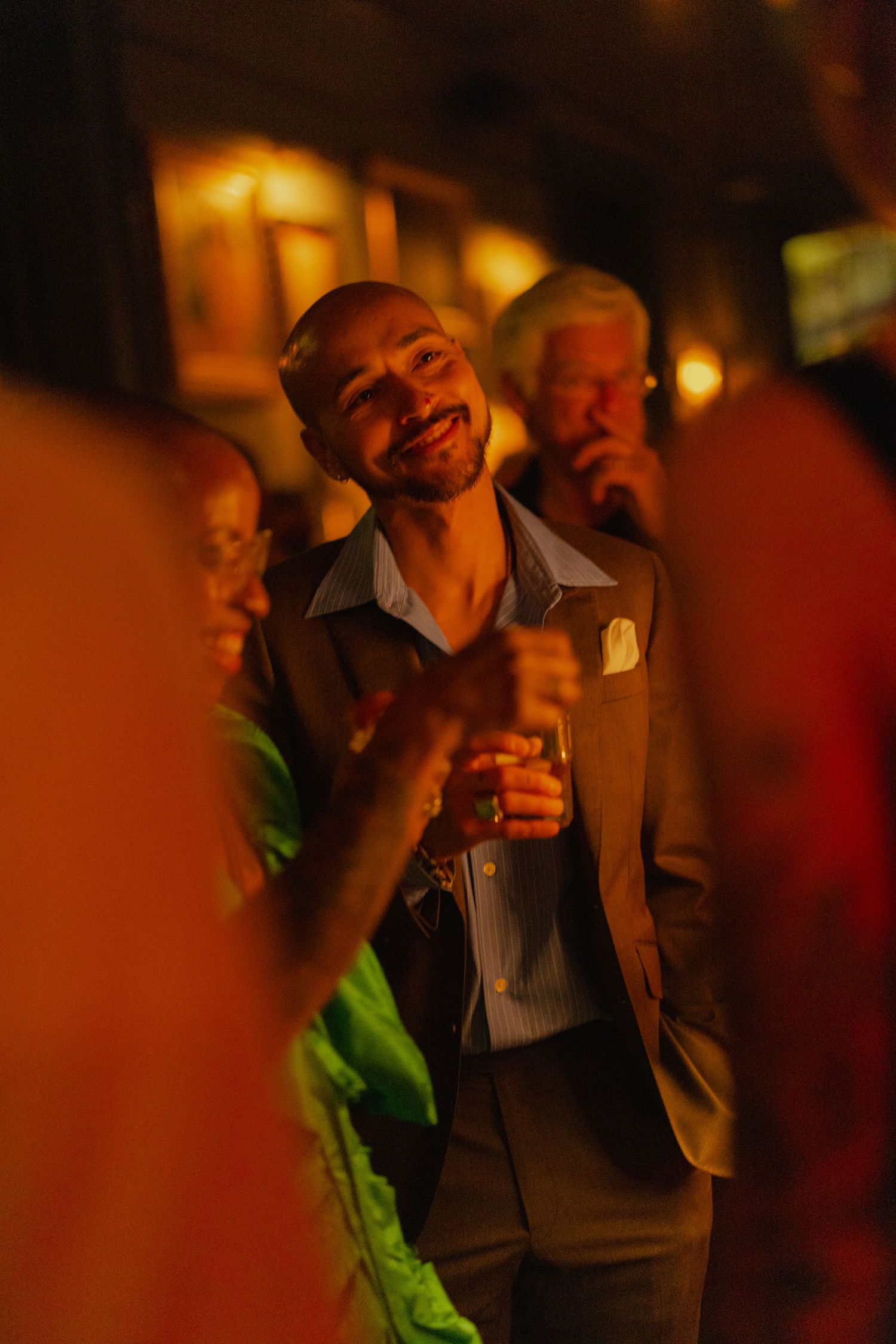 Smiling bald man in brown suit holds drink at warmly lit evening social gathering.