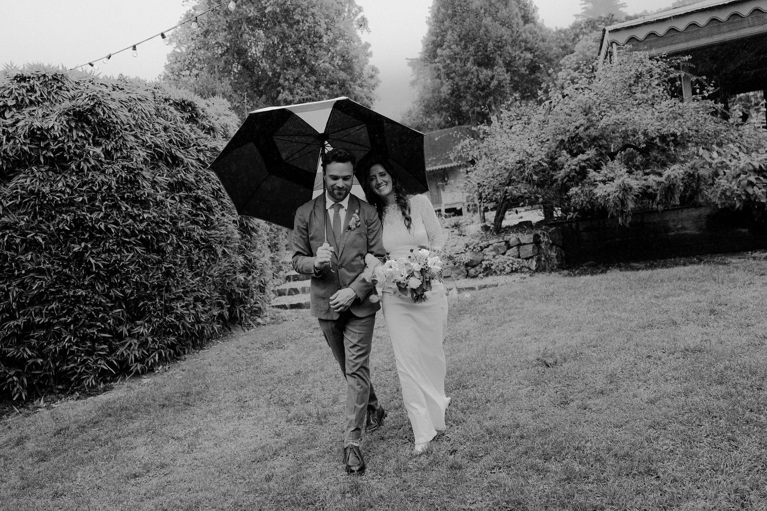 Bride and groom walking through lush garden in the rain sharing a black umbrella on their wedding day.