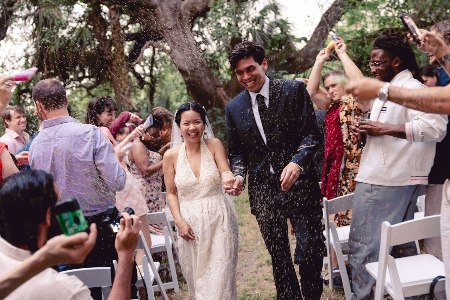 Joyful newlyweds walk down outdoor aisle as guests toss confetti at garden wedding ceremony.