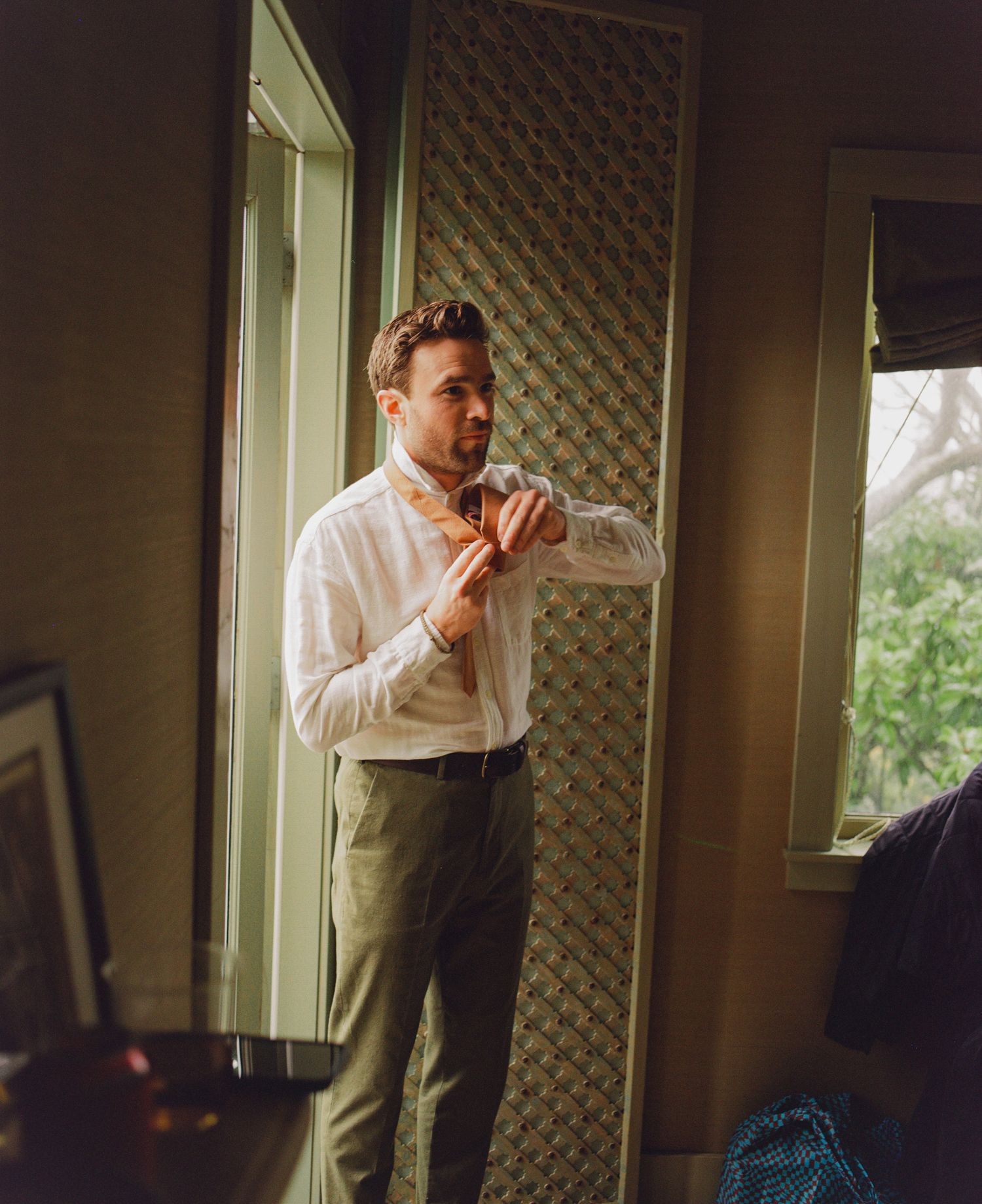 Groom in white shirt and olive trousers adjusting his tie by a window before the wedding ceremony.