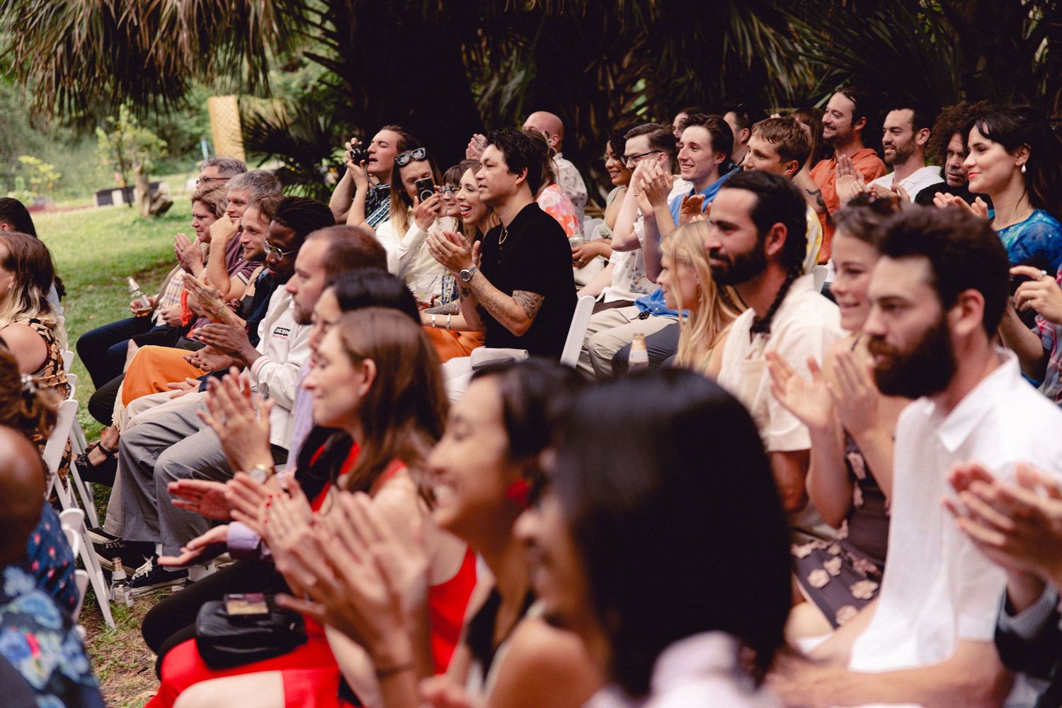 Wedding guests clap and celebrate during outdoor ceremony seated in sunny garden venue.