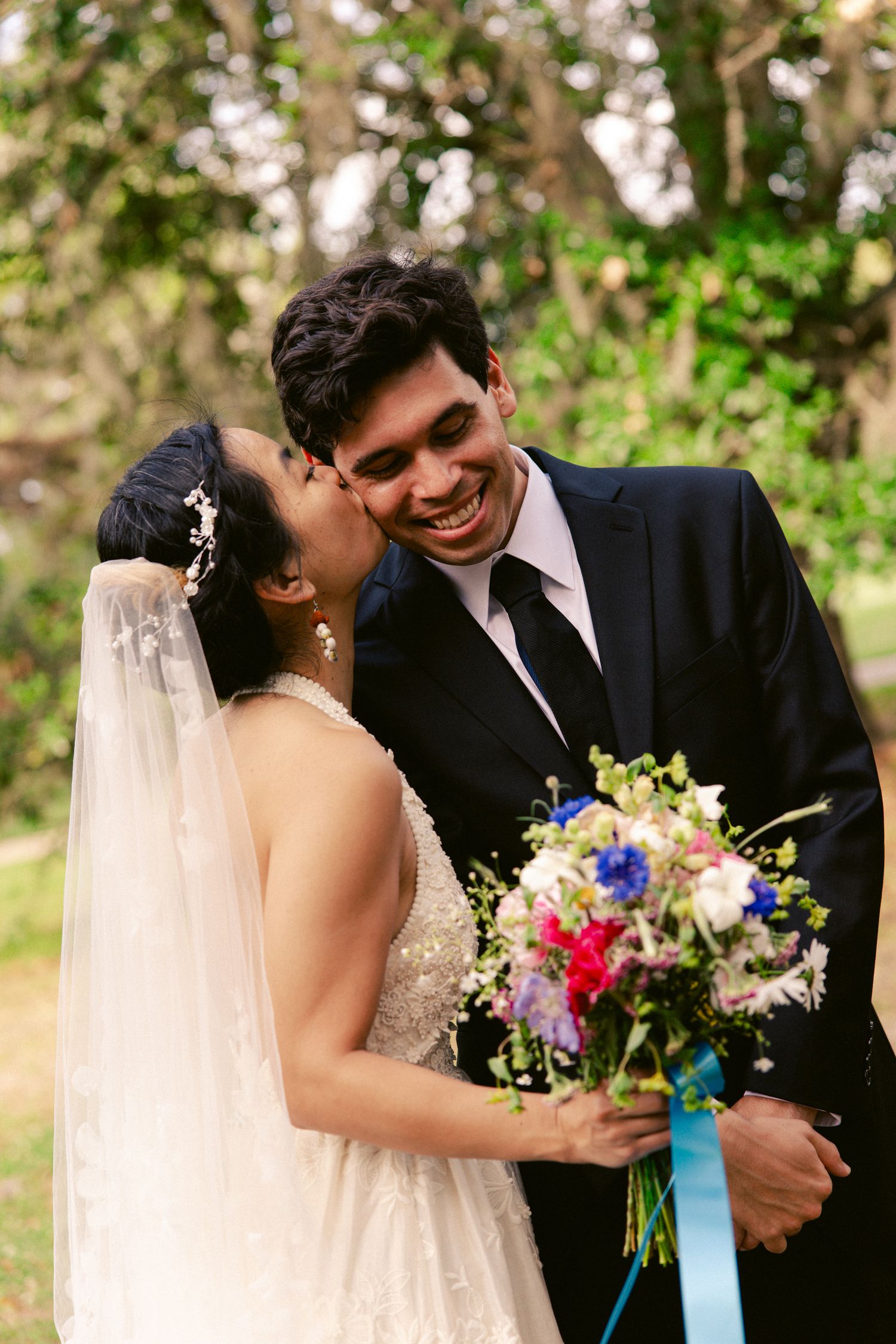 Bride kisses groom on cheek as he smiles joyfully, holding colorful wedding bouquet outdoors.