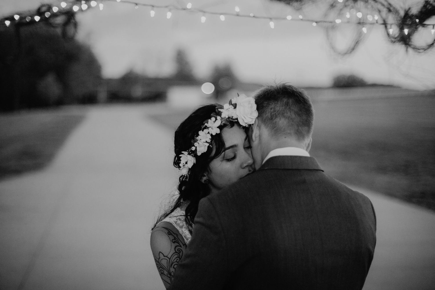 Bride with floral crown leans into groom under twinkling string lights at outdoor wedding.