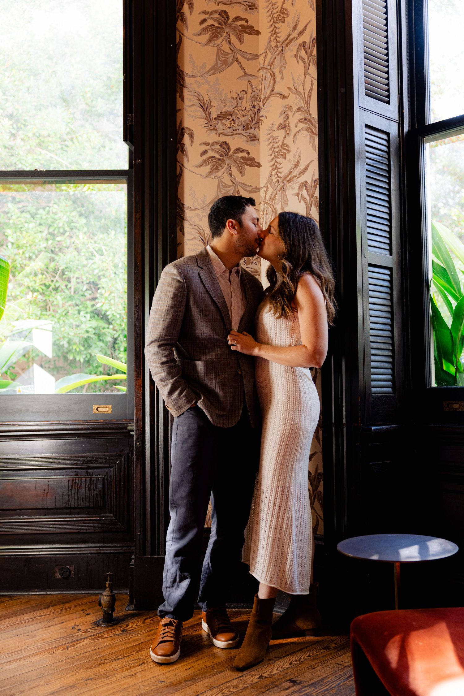 Couple sharing a romantic kiss by a window with floral wallpaper in an elegant interior setting.