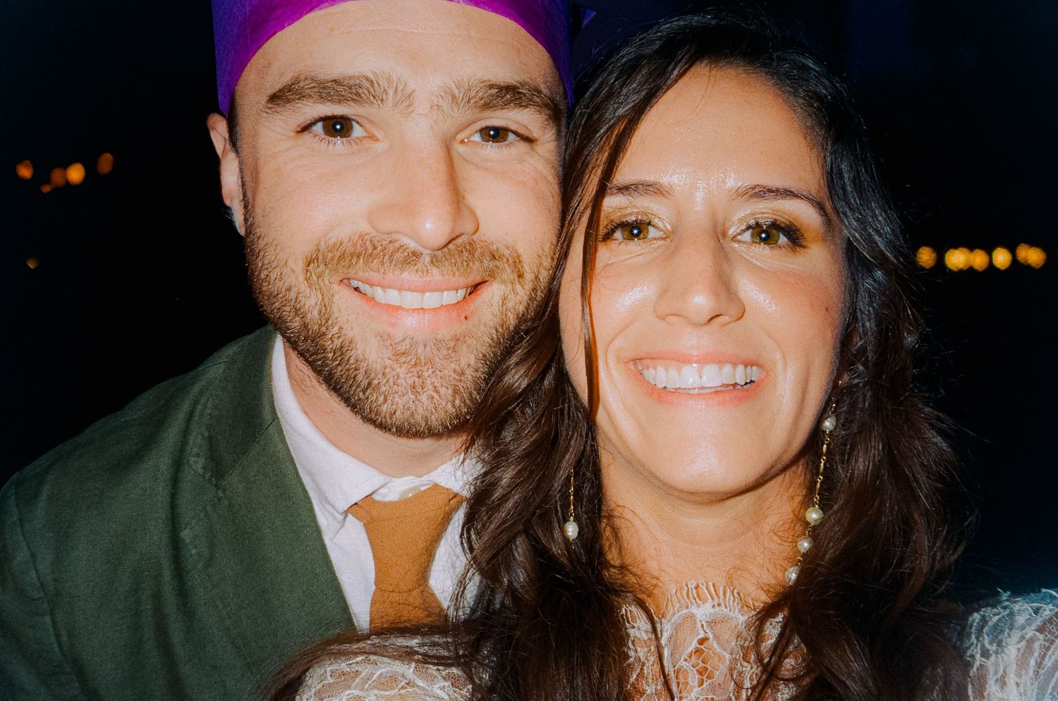 Smiling couple poses for a close-up selfie at a festive wedding reception with colorful lighting.