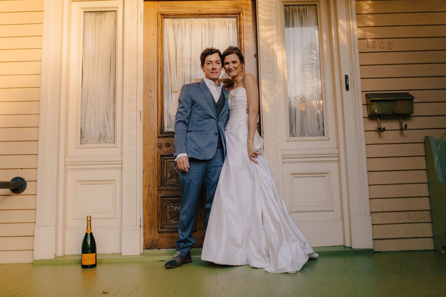 Bride and groom pose together in front of a white door with a champagne bottle on the porch floor.