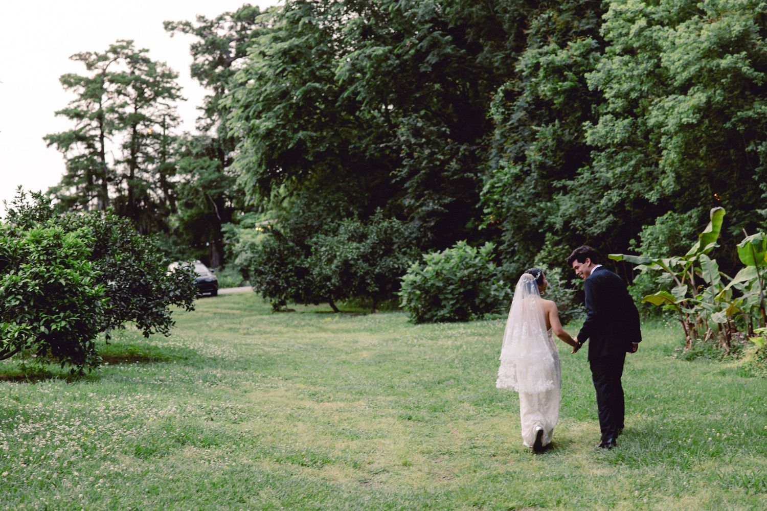 Bride in white gown and veil walks hand in hand with groom across lush green lawn at outdoor wedding.