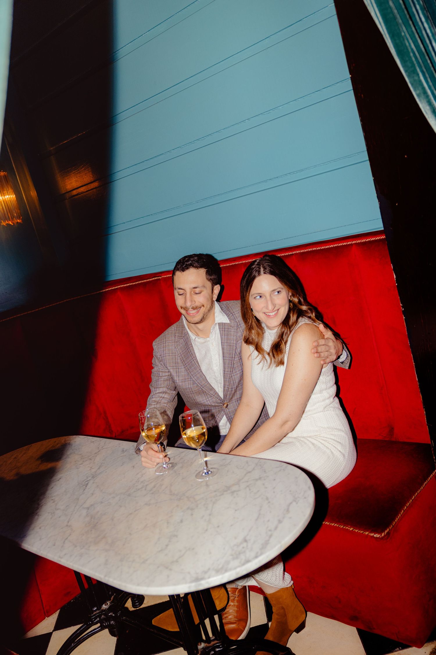 Couple smiling and holding champagne glasses at a marble table in a red velvet booth at an upscale restaurant.