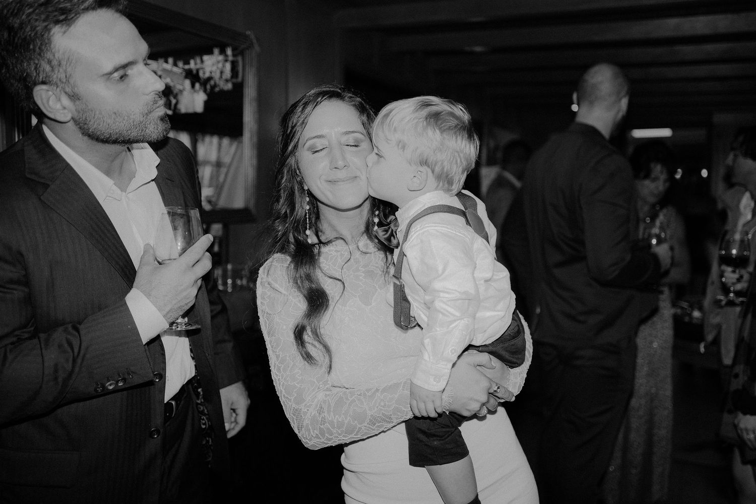 Black and white photo of a woman holding a young child who kisses her cheek at a wedding reception.