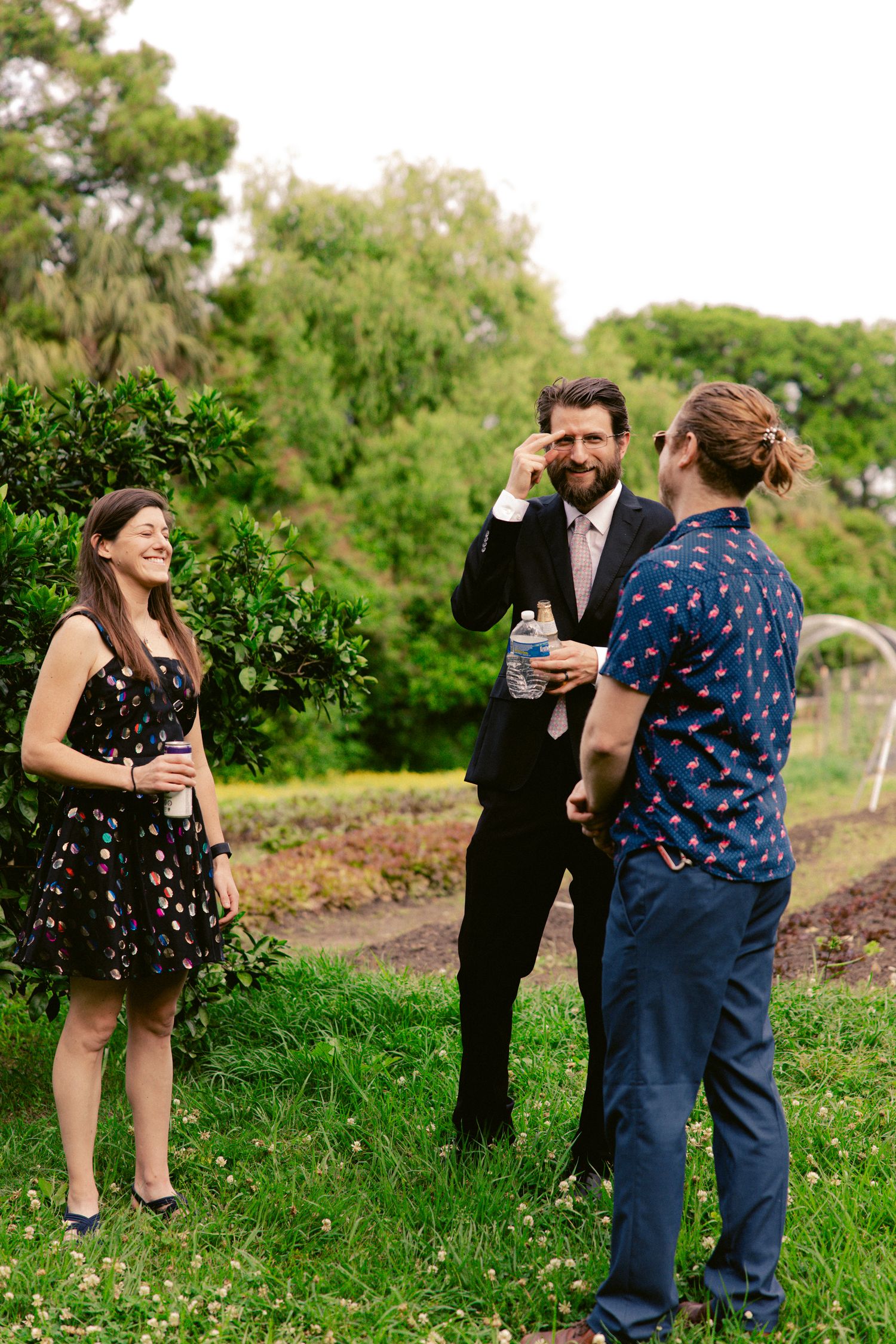 Three wedding guests in a garden setting enjoy drinks and conversation beside lush green shrubs.