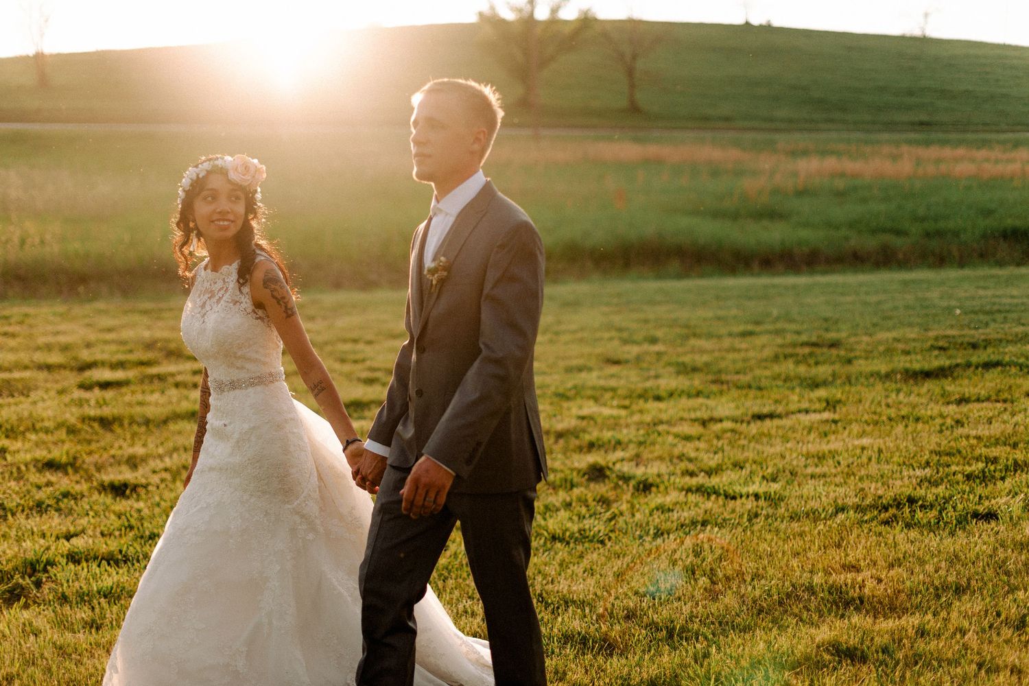 Bride in lace gown and groom in gray suit walk hand-in-hand through a glowing sunset field.