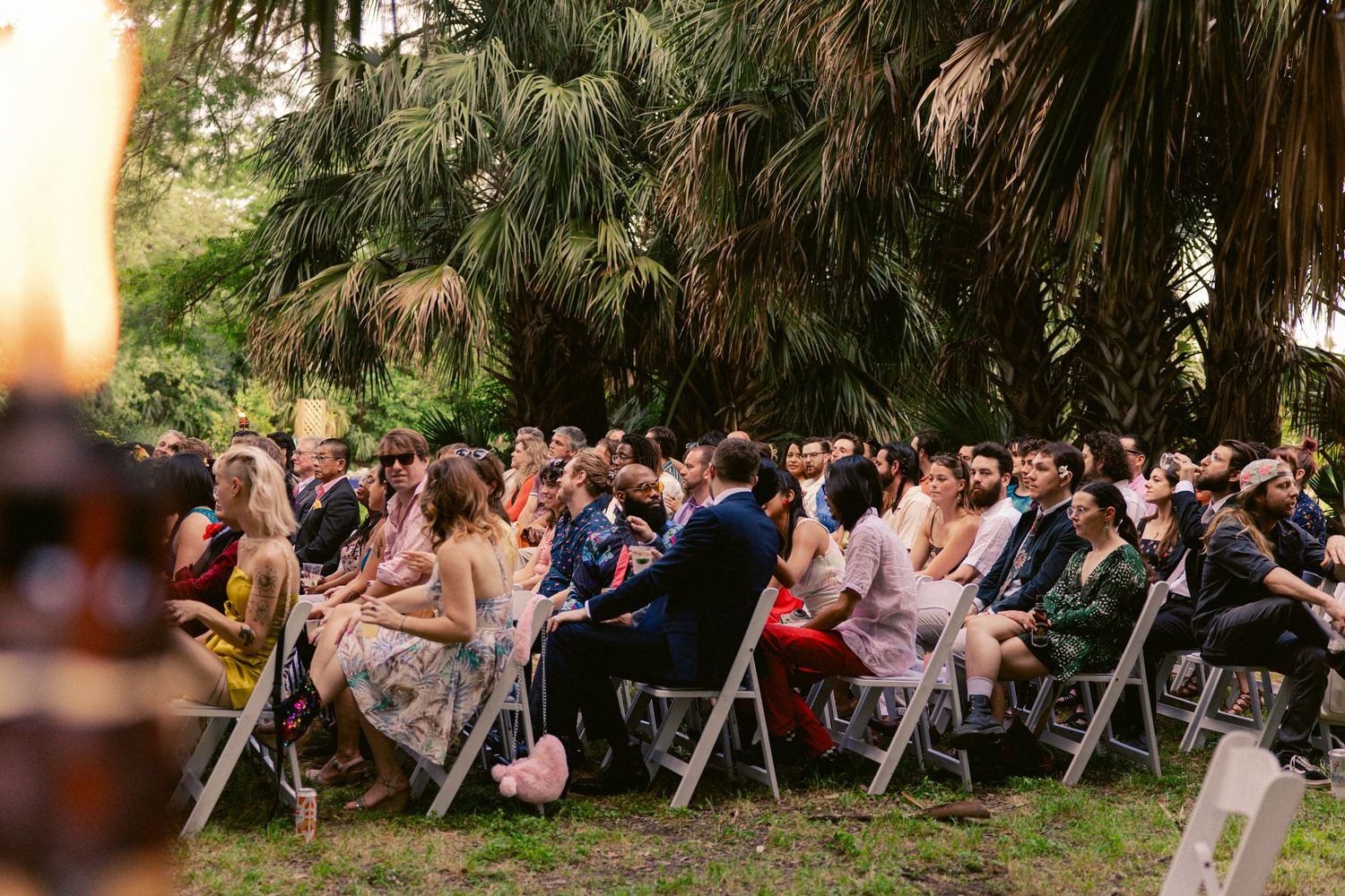 Large group of wedding guests seated in white folding chairs at a lush outdoor garden ceremony.