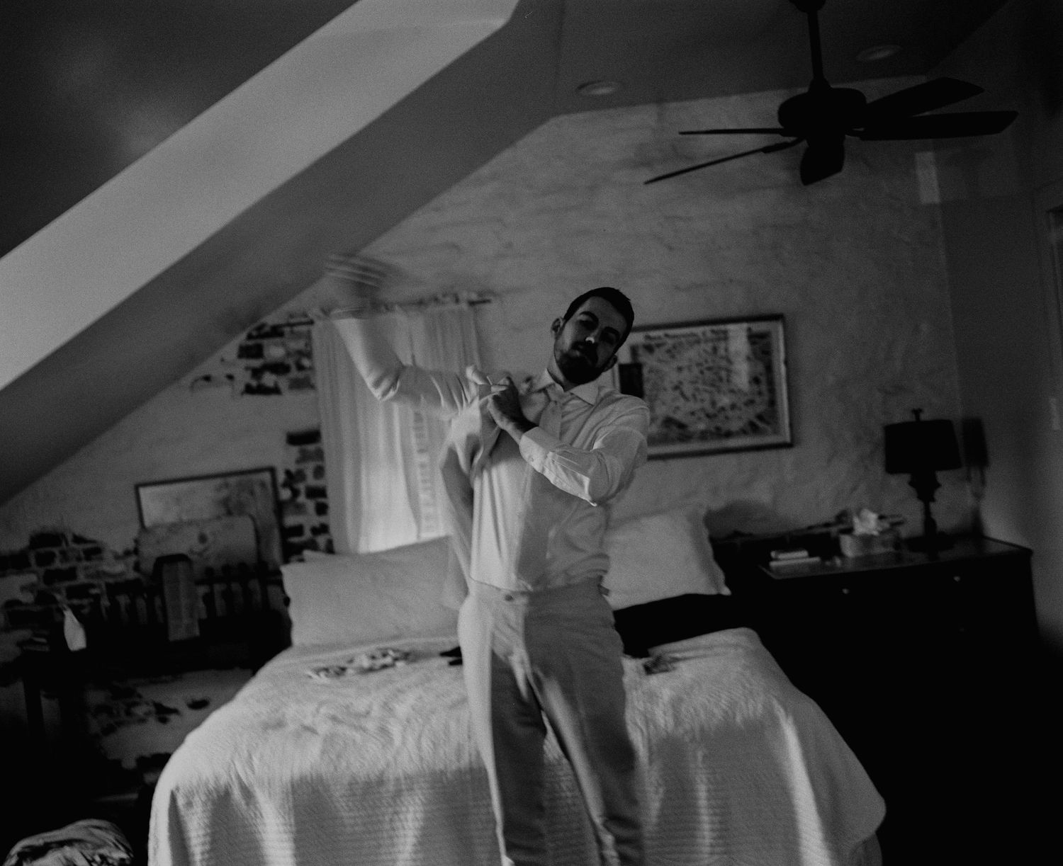 Man adjusting his tie in a bedroom with sloped ceiling and ceiling fan, black and white wedding preparation photo.