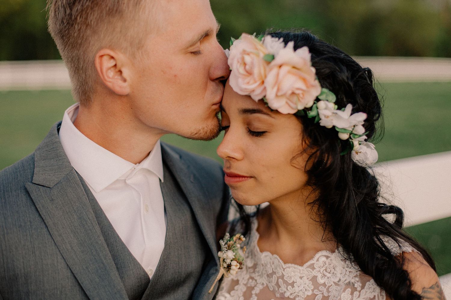 Groom in gray suit tenderly kisses bride wearing a pink floral crown at an outdoor wedding.