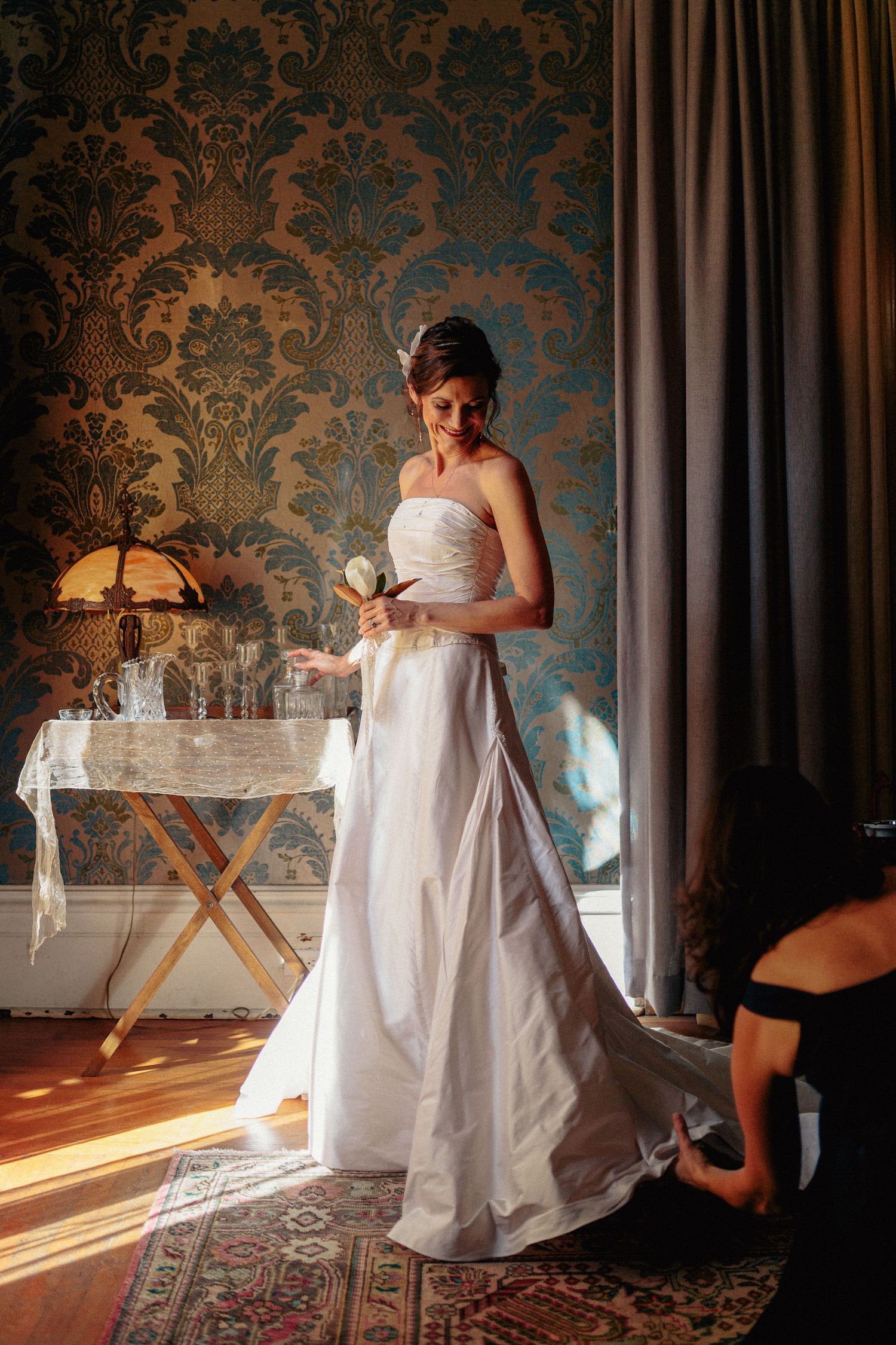 Bride in strapless white gown smiles as bridesmaid adjusts her train in ornate wallpapered room.