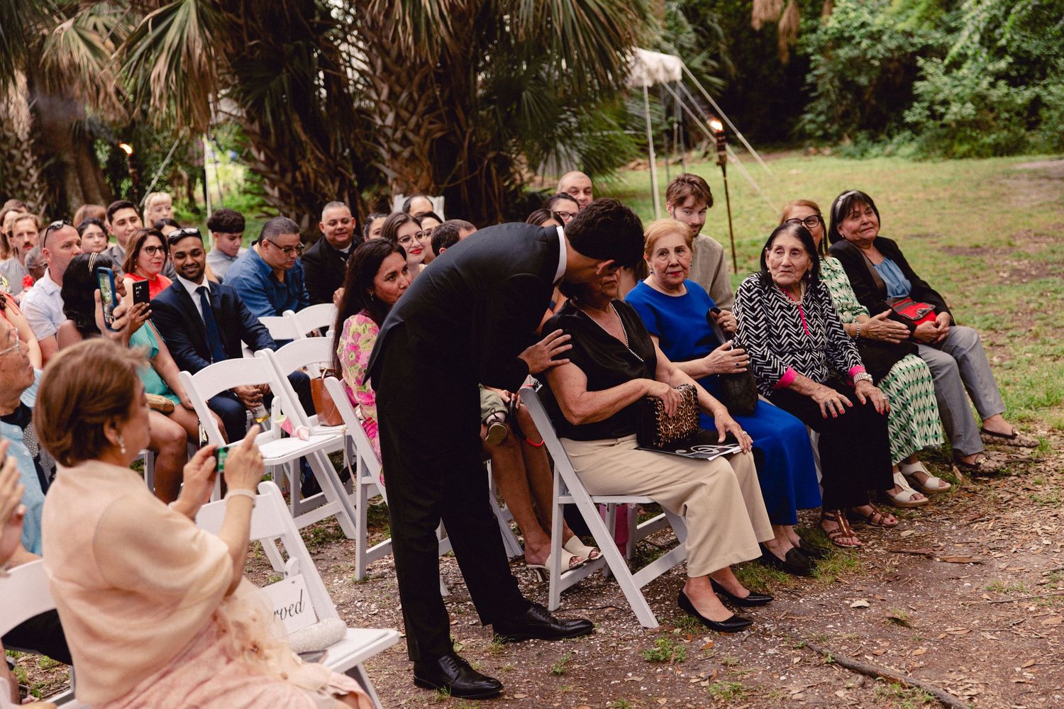 Groom in black suit greets seated wedding guests along the aisle before the outdoor ceremony begins.