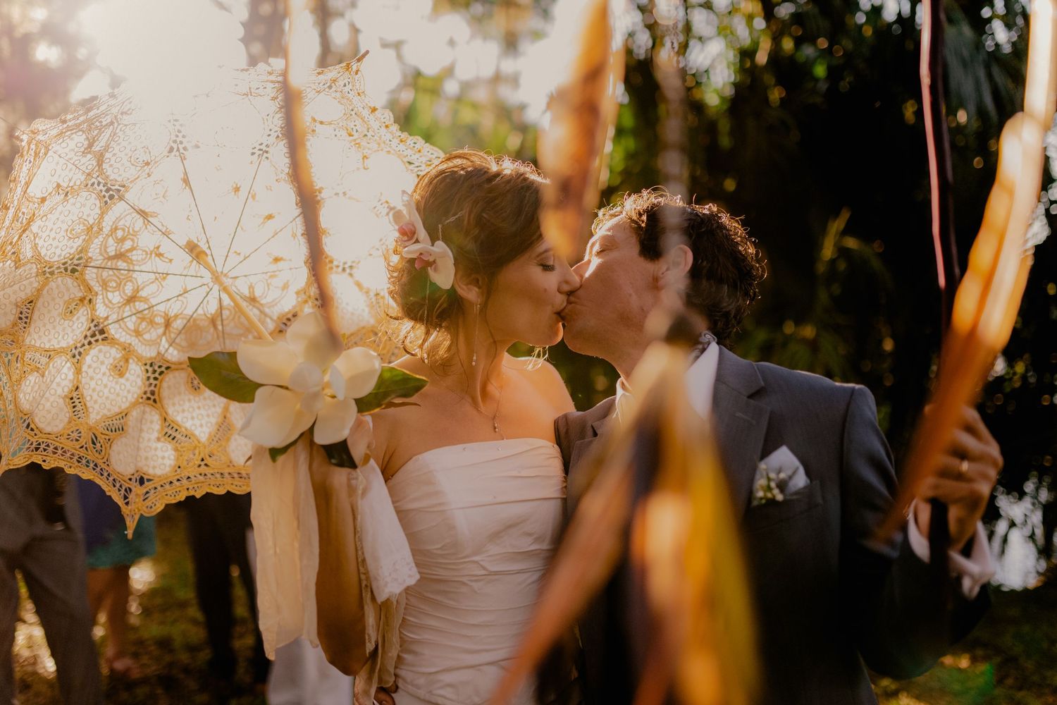 Newlyweds share first kiss under lace parasol at golden hour outdoor wedding ceremony.