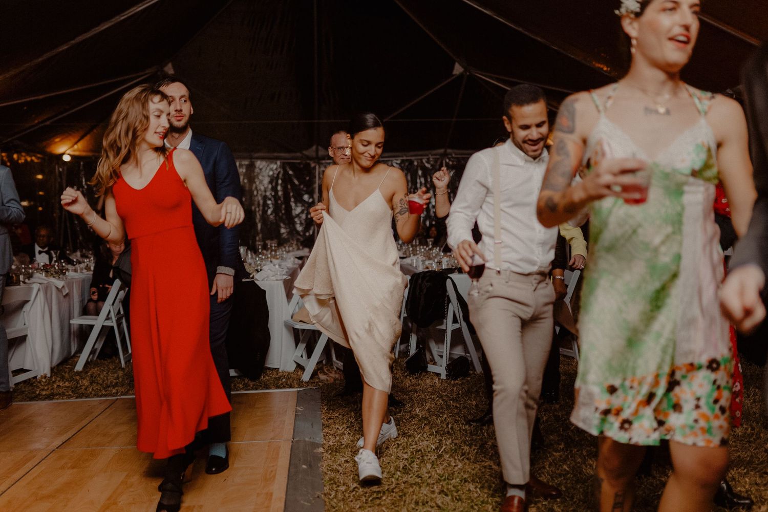 Wedding guests dancing joyfully in a tent reception, bride in white gown with colorful-dressed friends.