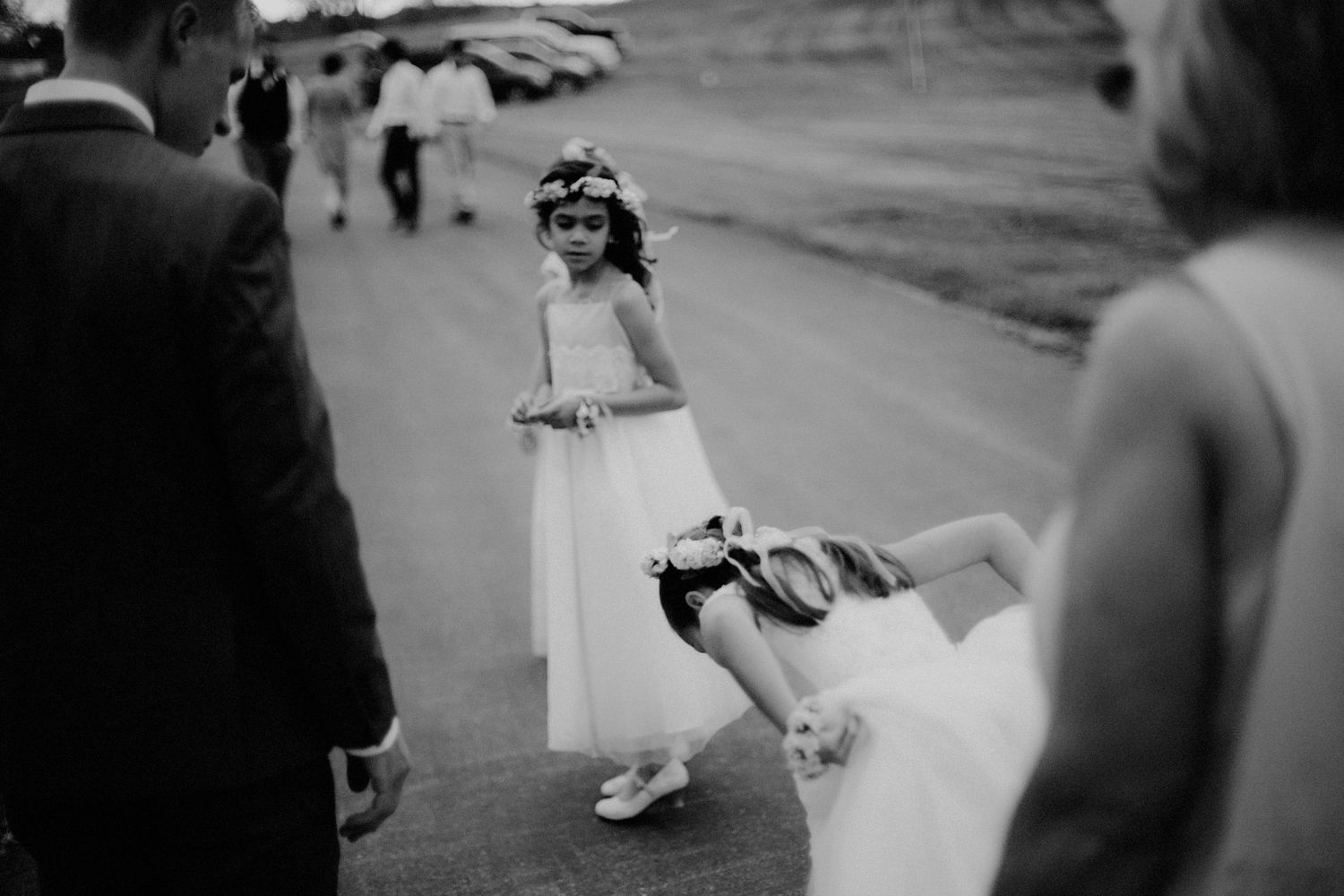 Flower girl in white dress playfully bending during outdoor wedding procession.