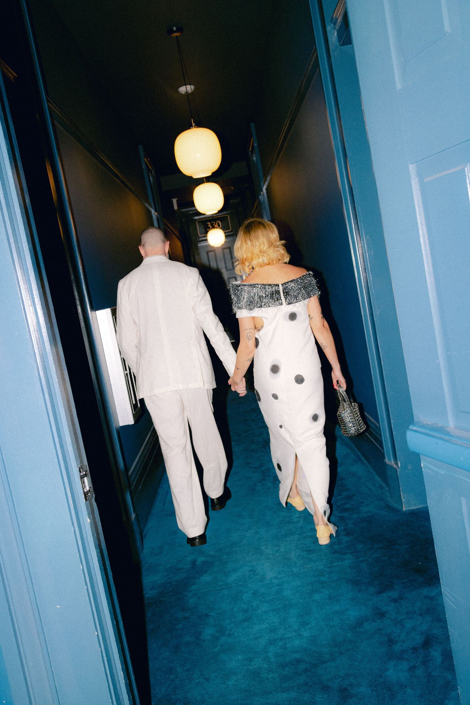 Newlywed couple walks hand-in-hand down vibrant teal hotel corridor after wedding ceremony.