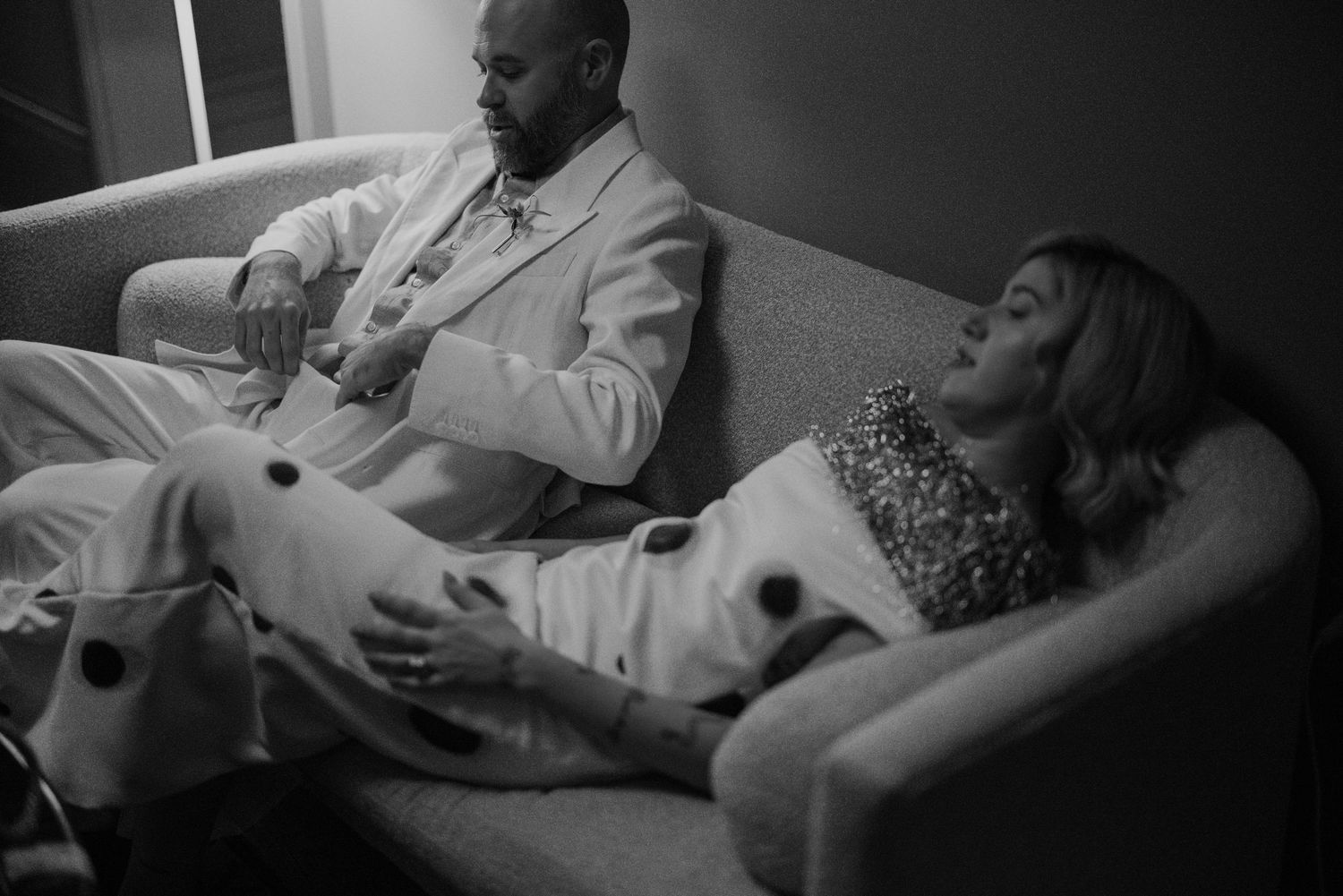 Black and white photo of bride and groom relaxing on sofa in wedding attire before ceremony.