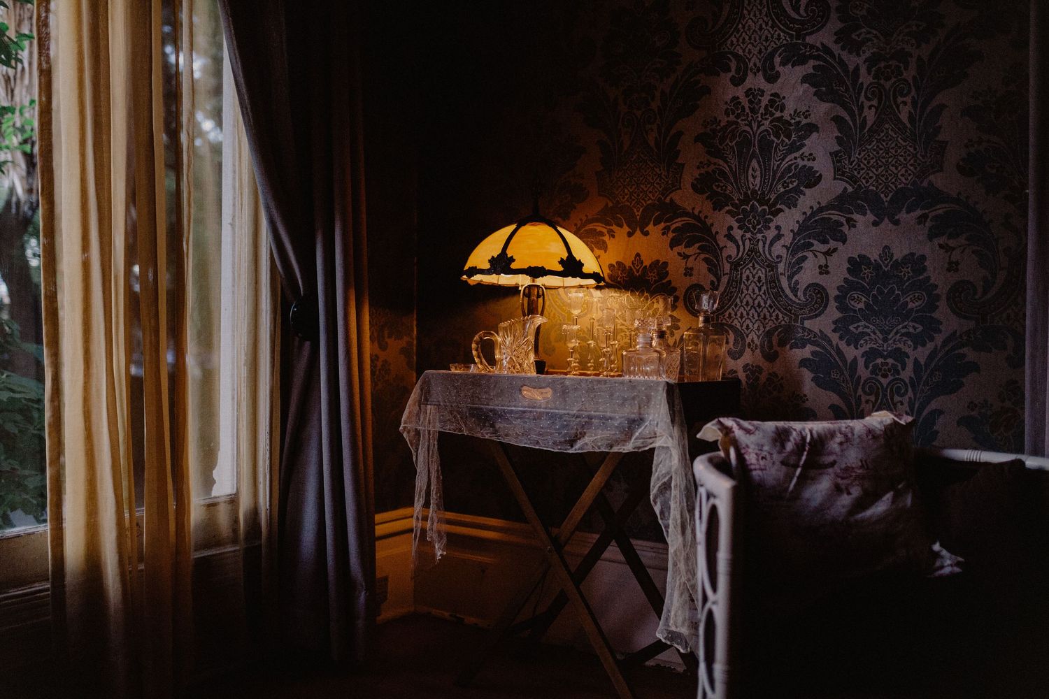 Vintage lamp glowing on an antique table beside damask wallpaper in a moody room.
