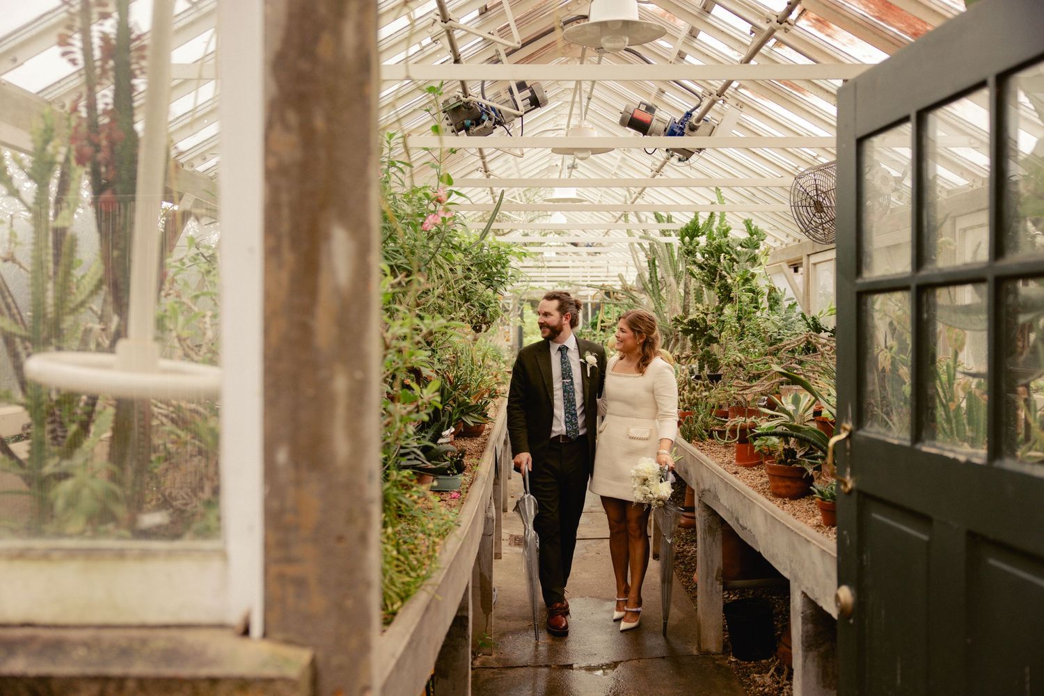Couple walking hand in hand through lush greenhouse during wedding photo session.