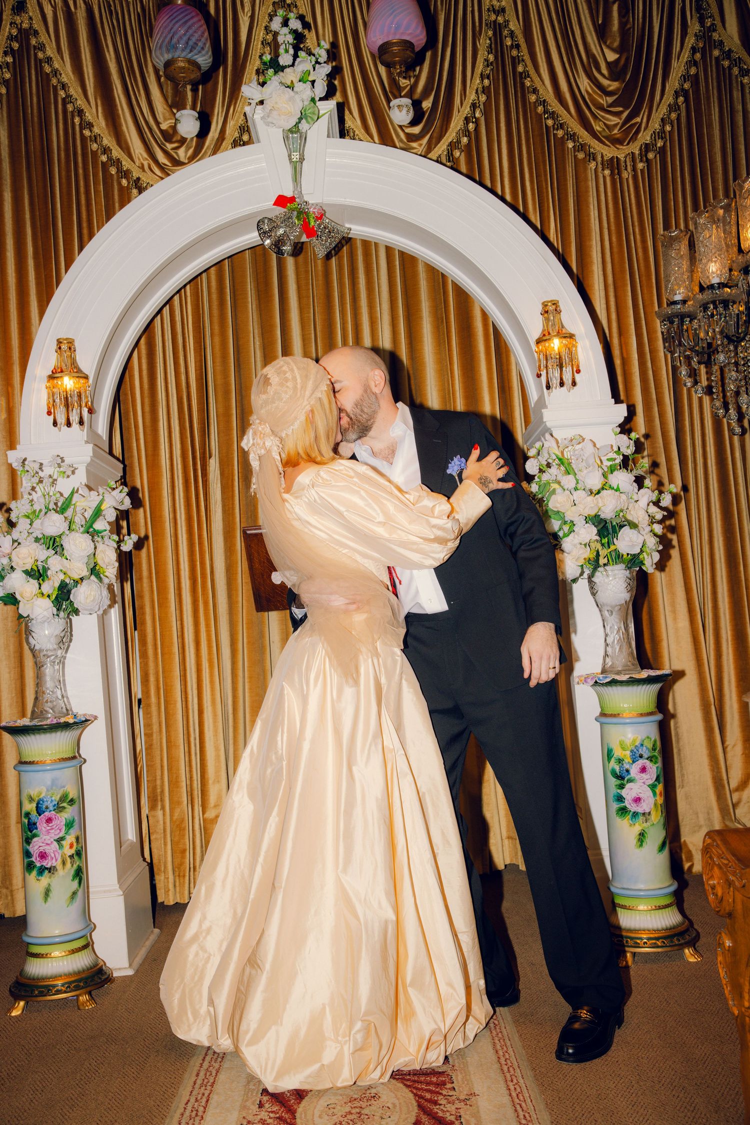 Bride and groom sharing first kiss under white floral arch with golden curtain backdrop.