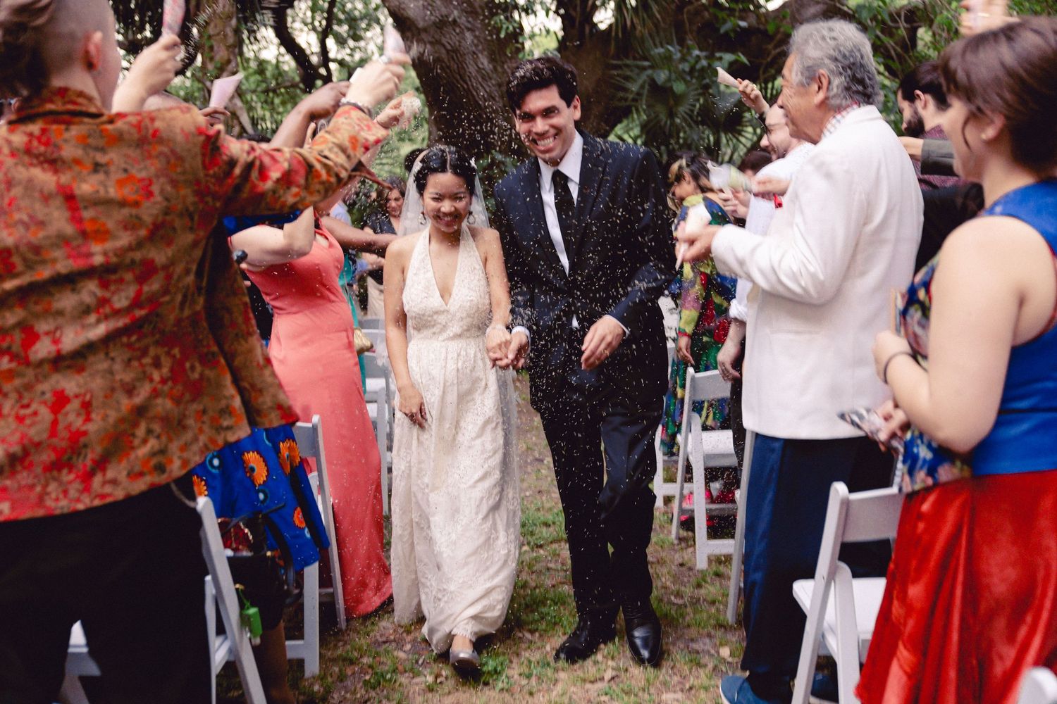 Guests shower a laughing bride and groom with confetti as they exit their outdoor woodland wedding.