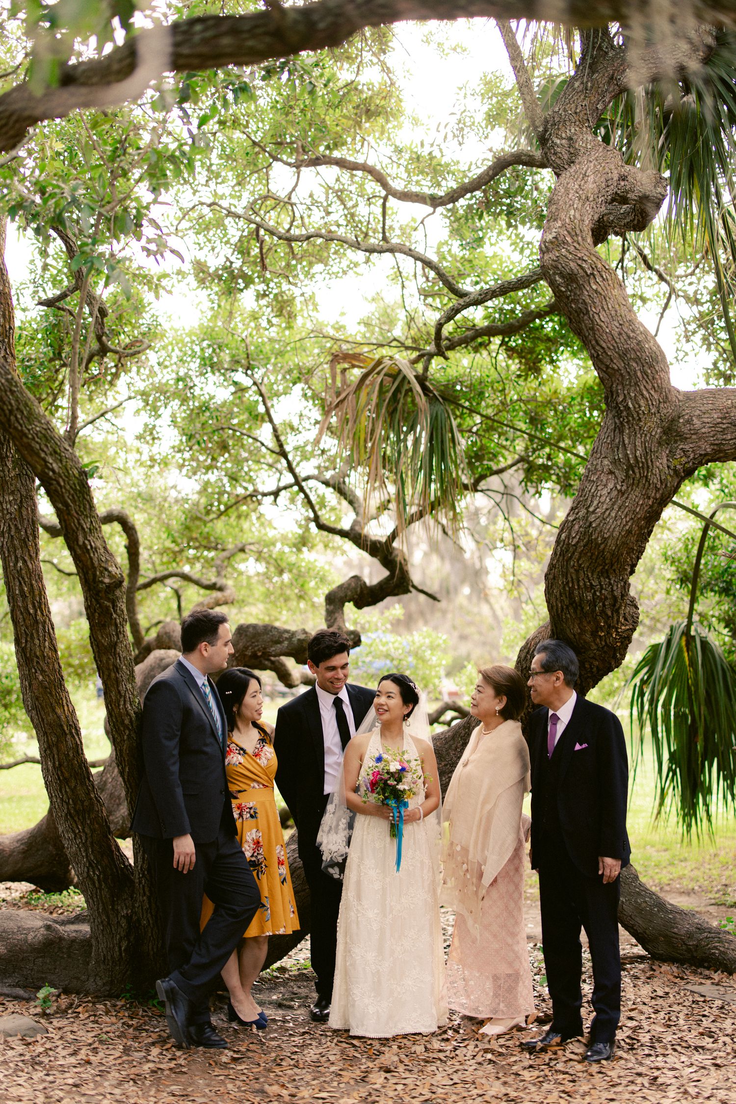 Wedding couple poses with family members under large oak trees in lush outdoor garden setting.
