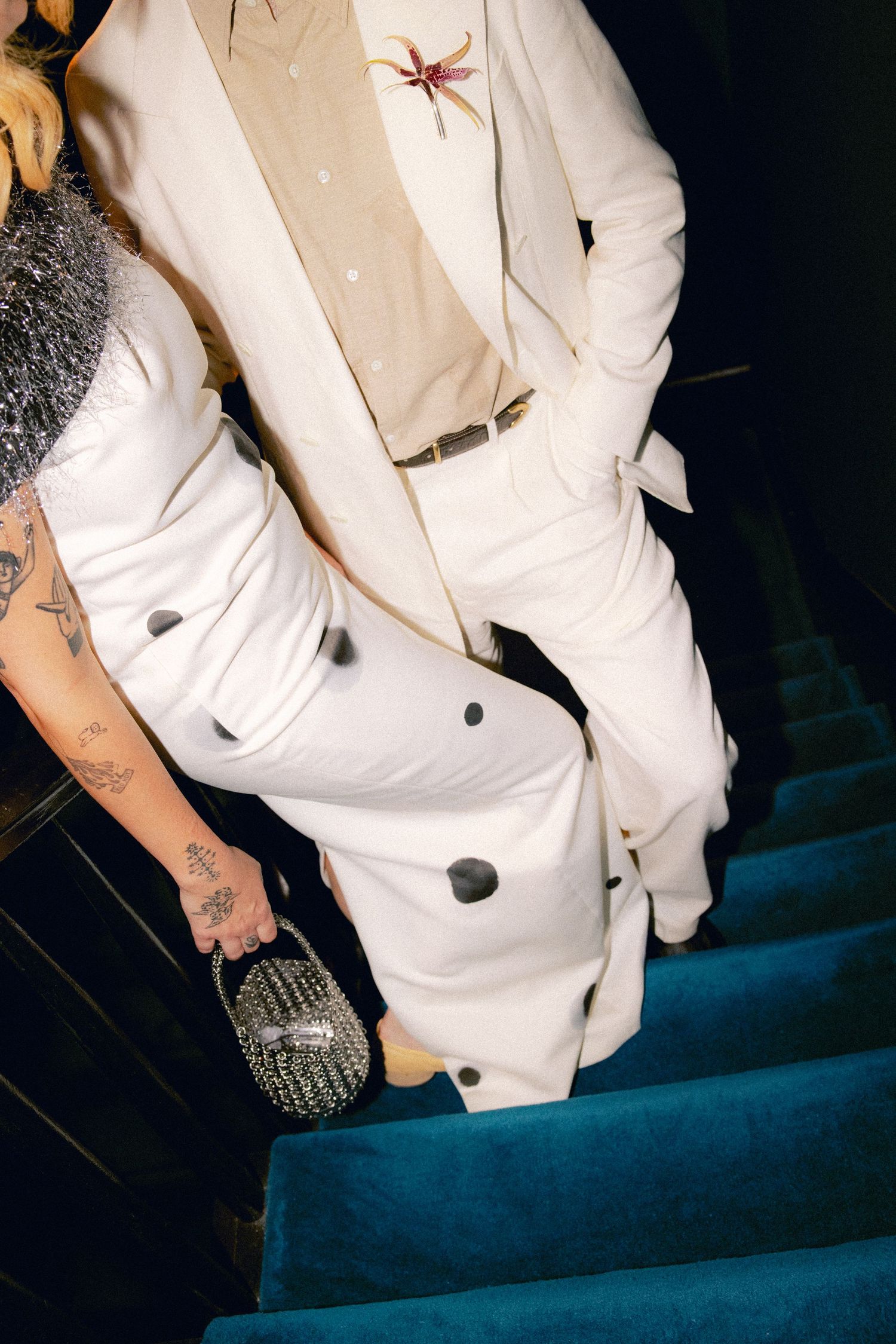 Person in white suit with floral brooch descends blue velvet stairs with tattooed companion.