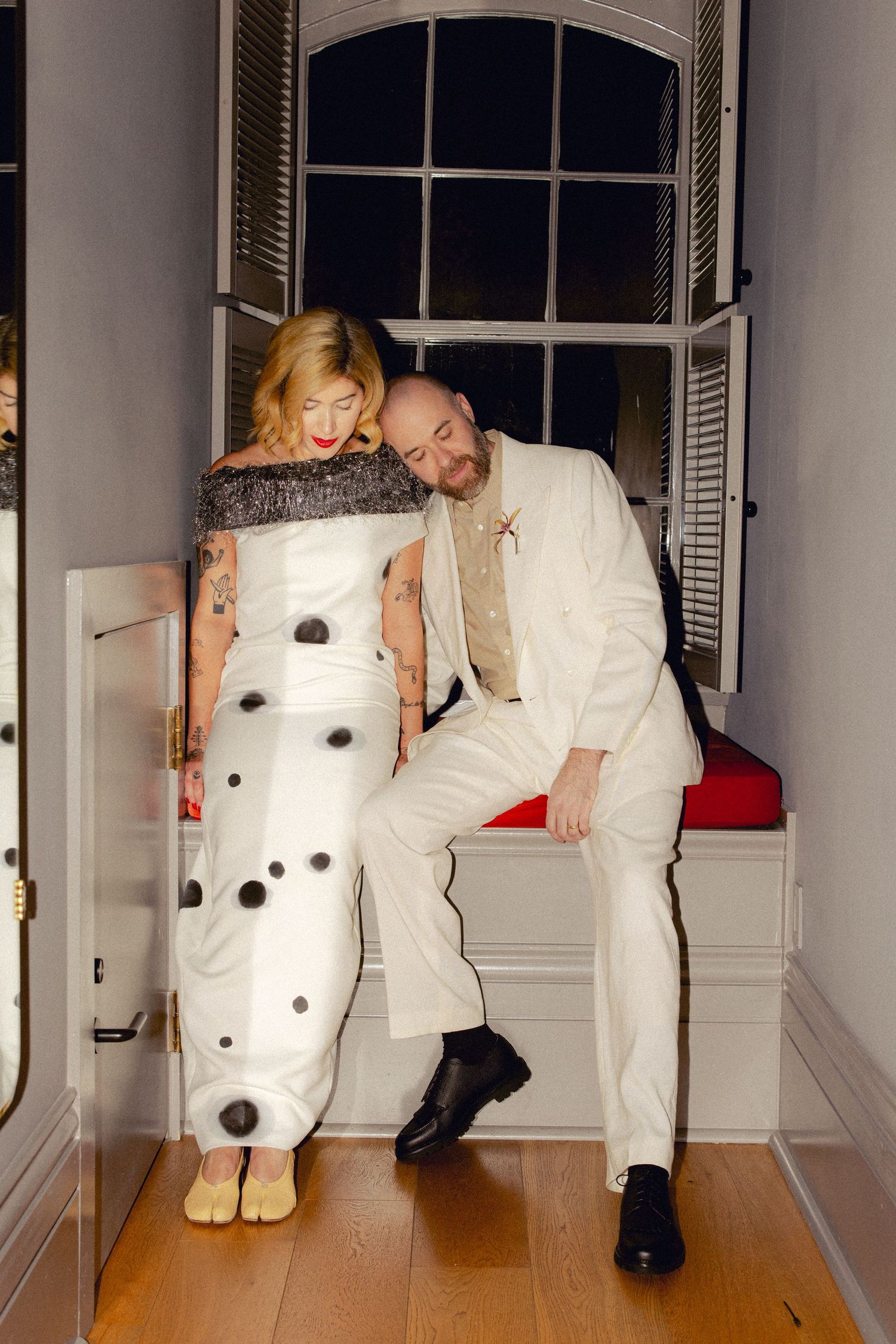 Bride in polka dot gown and groom in white suit posing playfully in a window alcove.