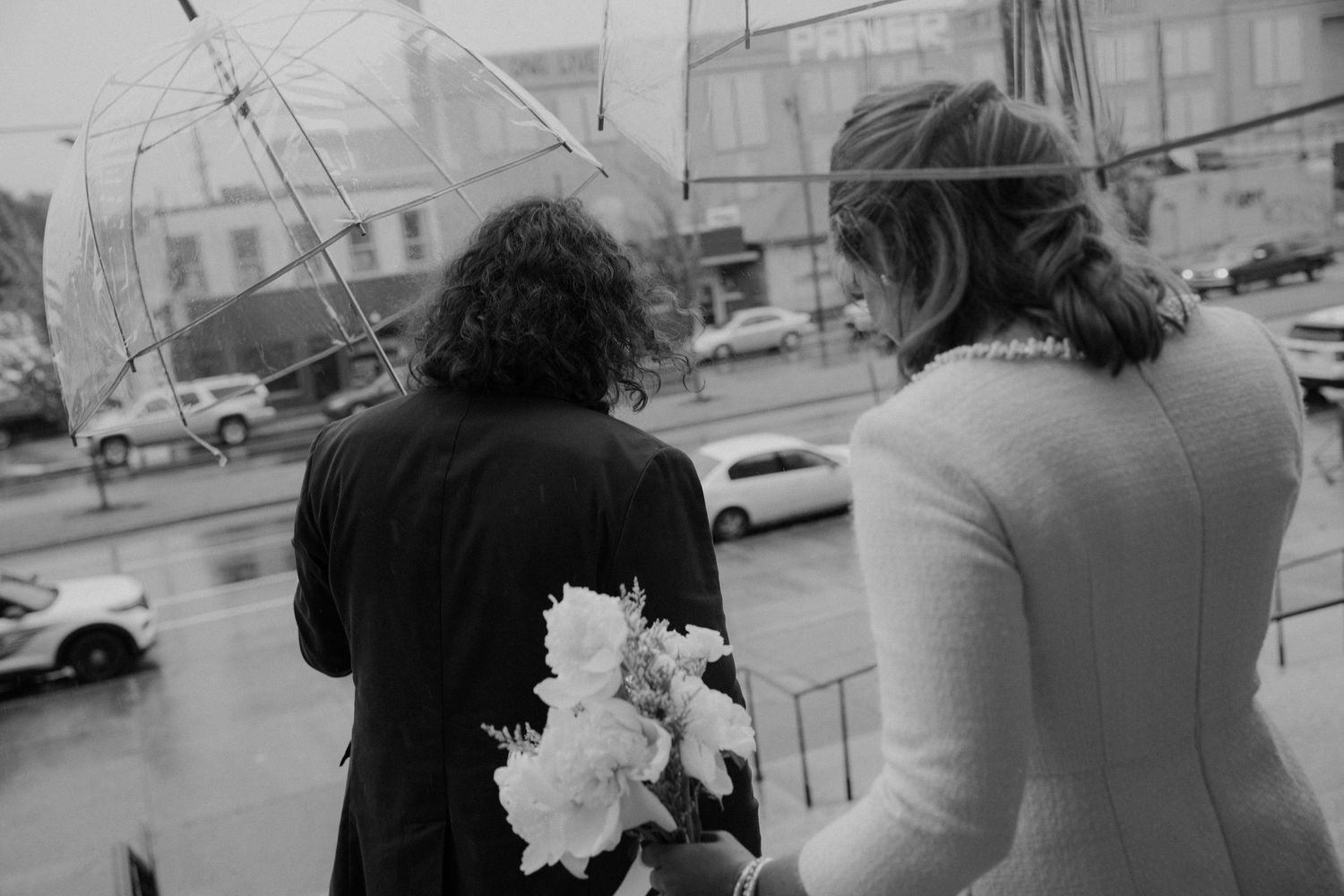 Black and white photo of bride and guest walking in rain with transparent umbrella.