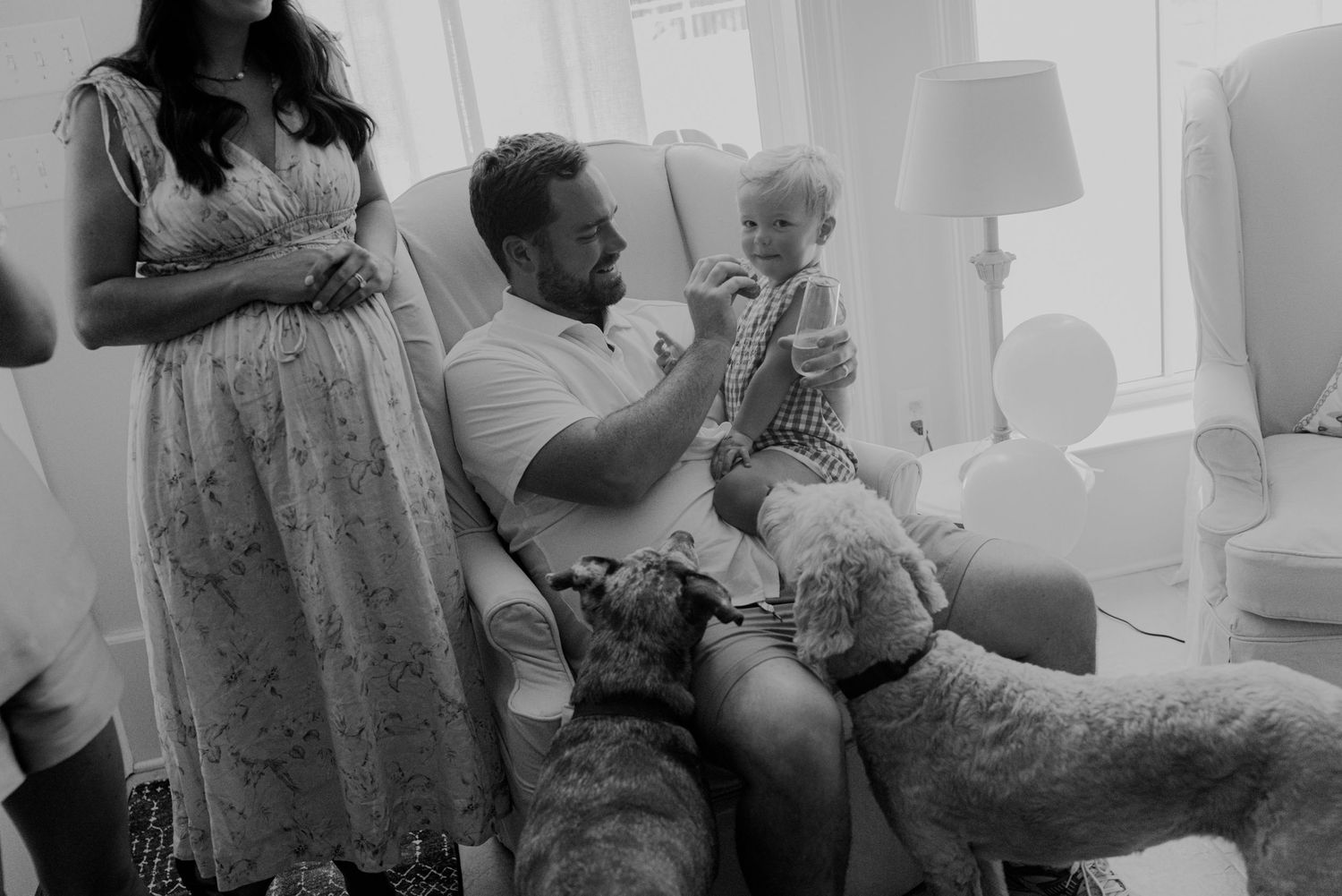 Father feeds baby in armchair while dog rests nearby in a cozy black and white family portrait.