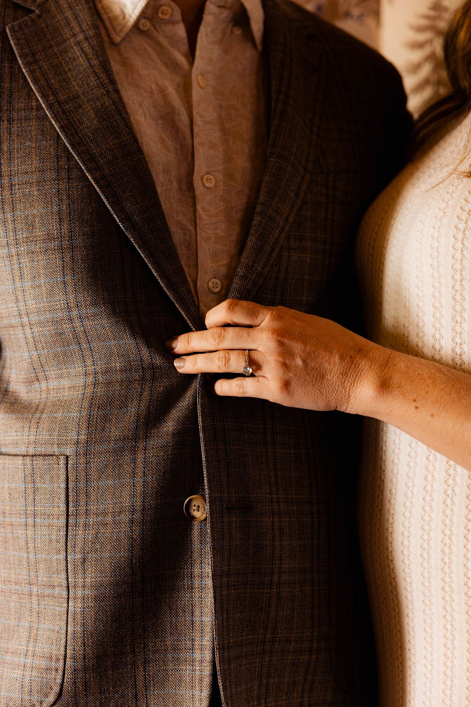 Close-up of bride's hand with engagement ring resting on groom's brown plaid suit jacket.