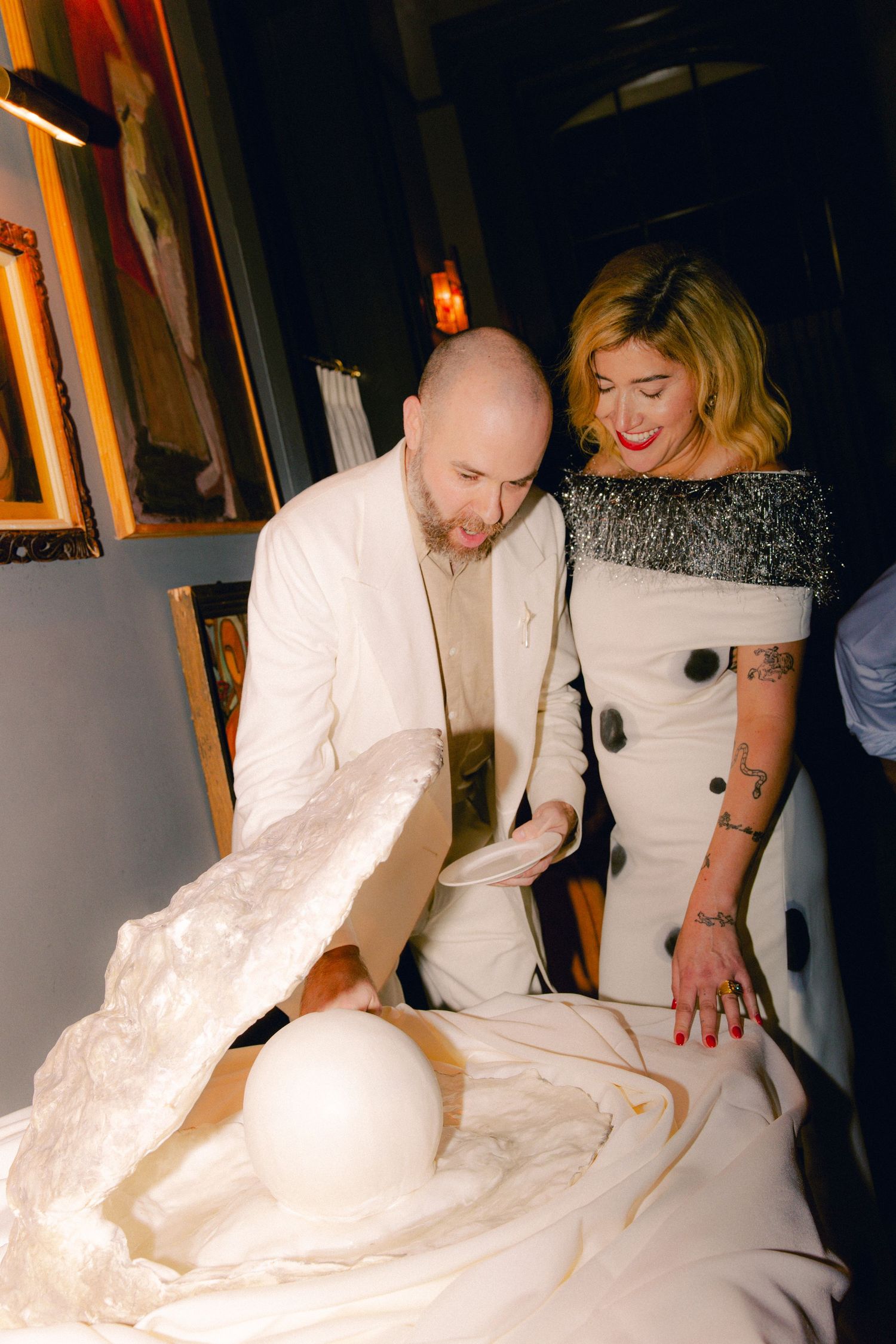Bald man in white suit and blonde woman examine white sculptural art installation indoors.