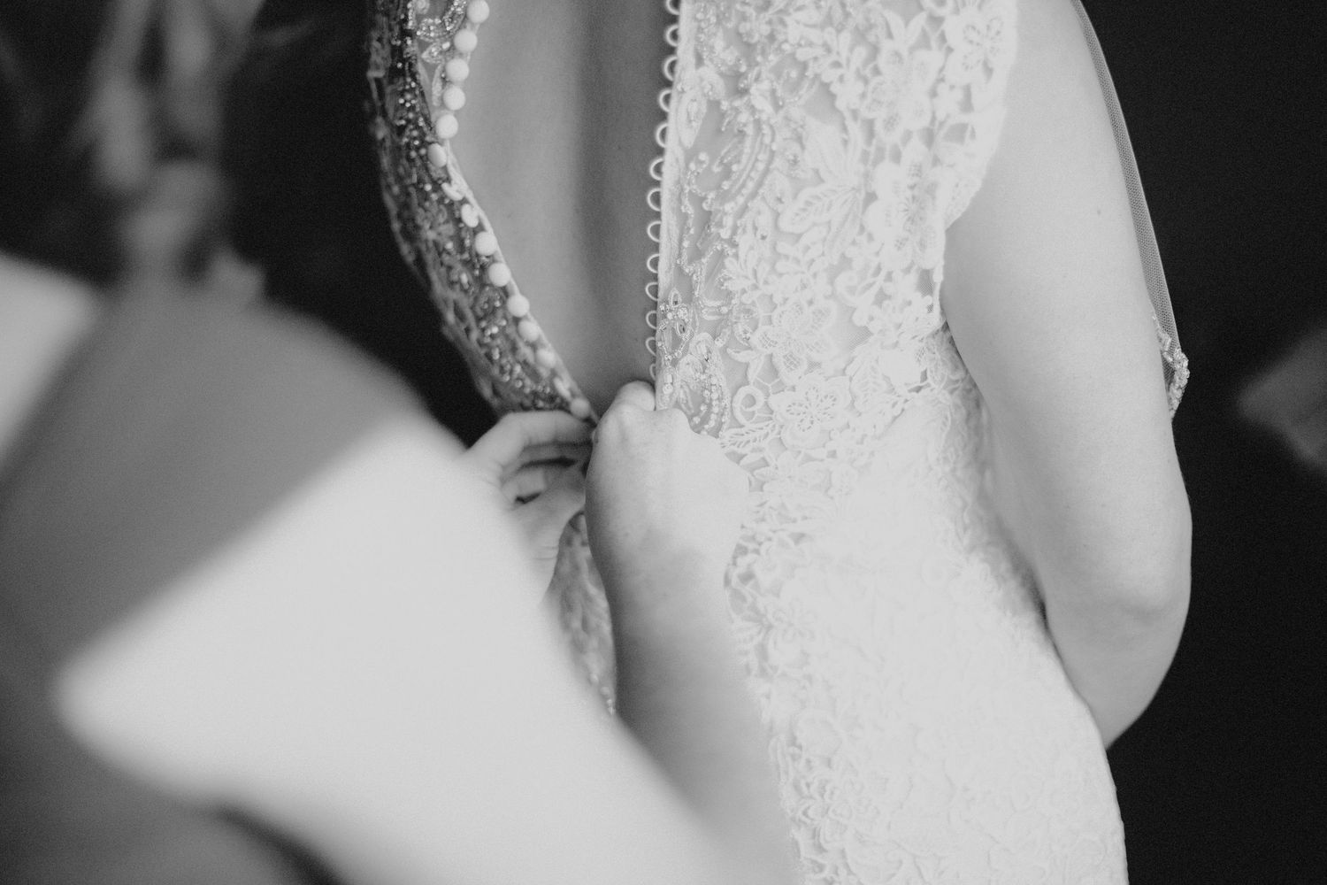 Close-up black and white photo of a bride having her lace wedding dress buttoned up the back.