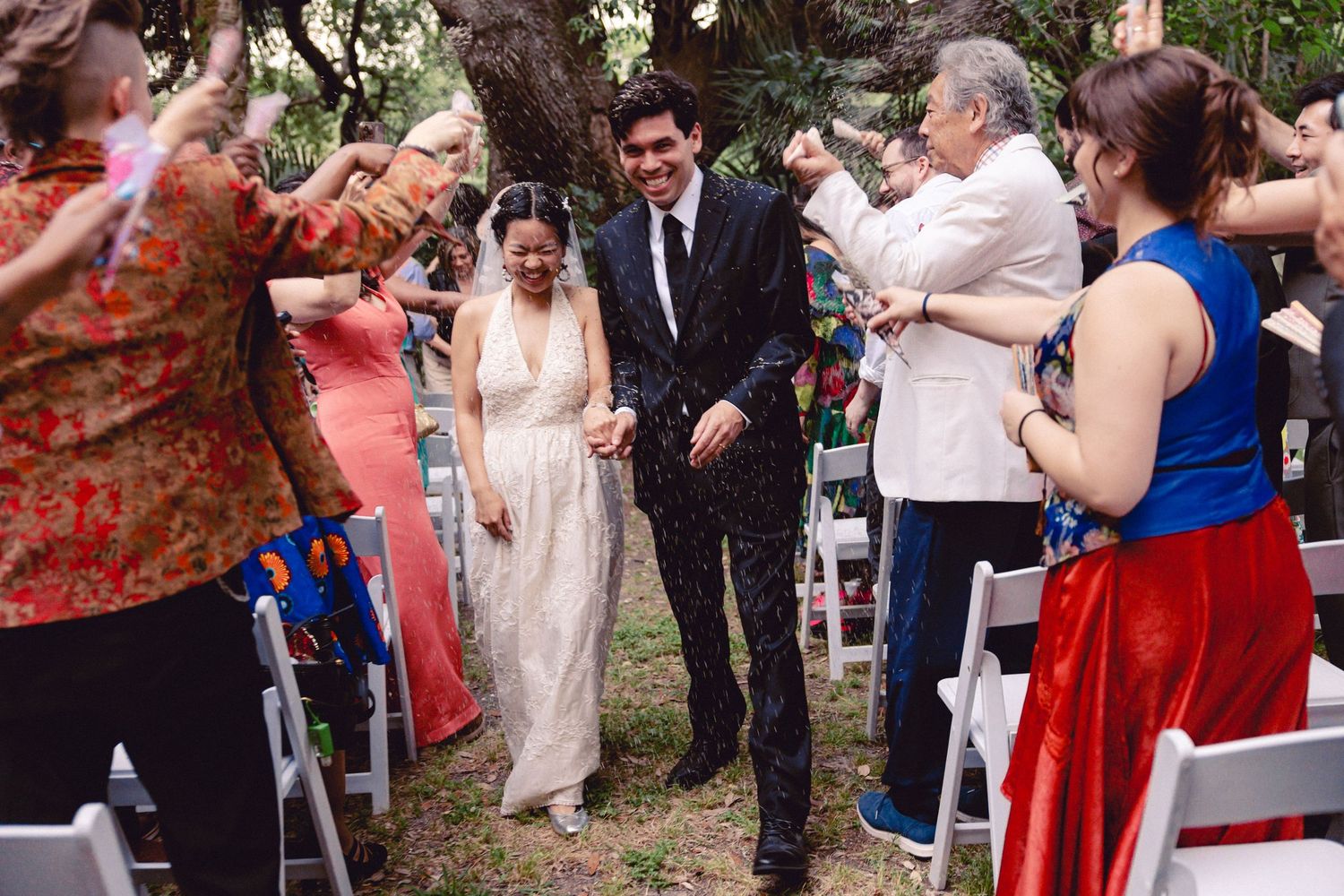 Bride and groom walk down aisle as guests celebrate with confetti at outdoor wedding ceremony.
