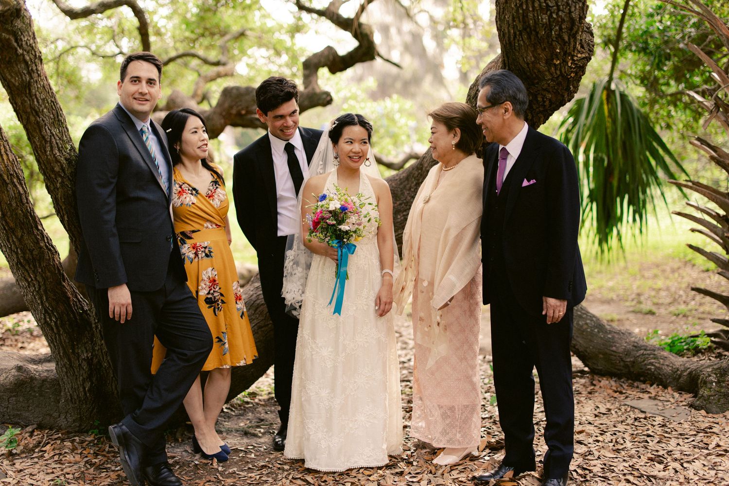 Bride and groom posing with family members under trees in an outdoor wedding portrait.
